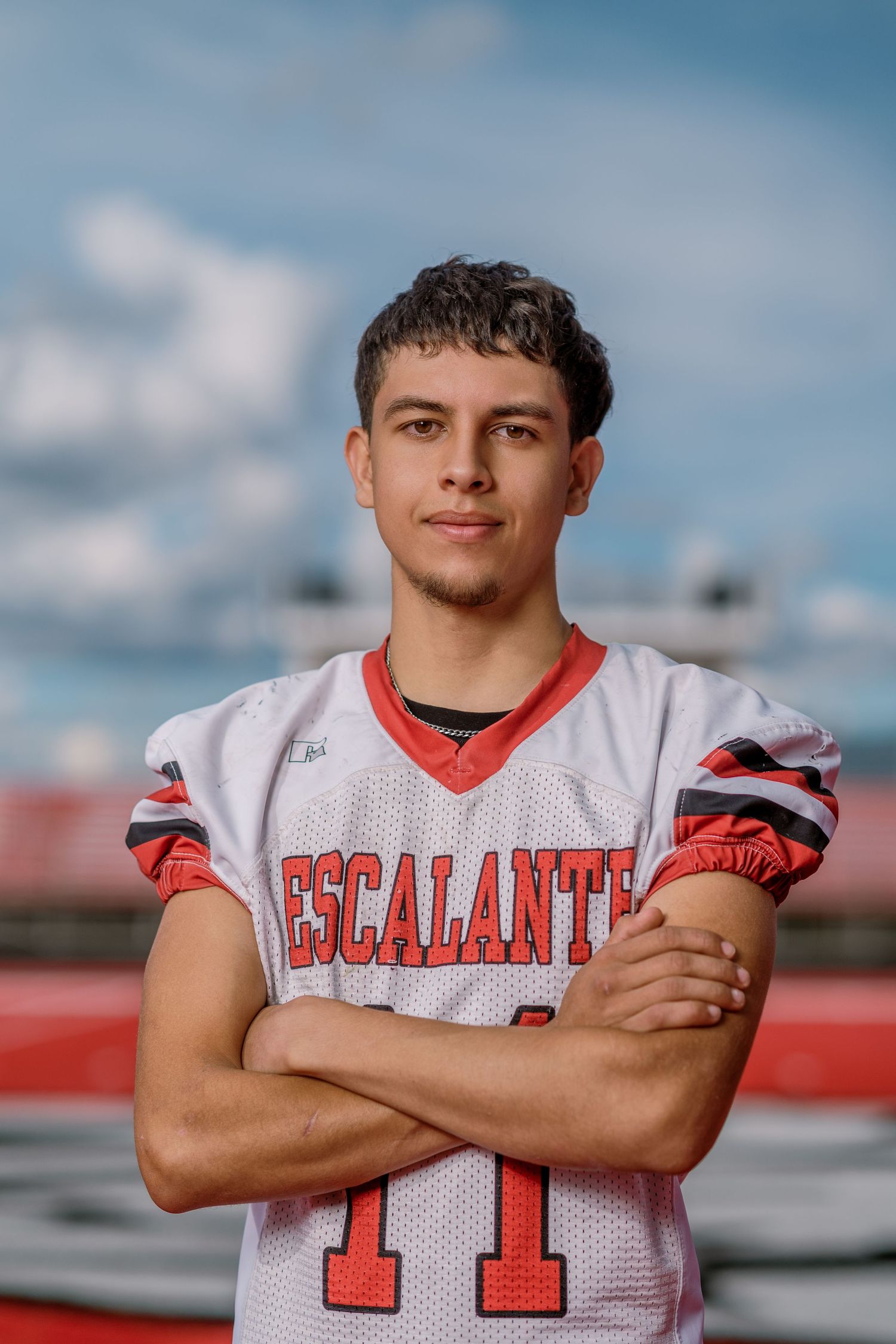 High school football player poses in white and red uniform against blue sky background.