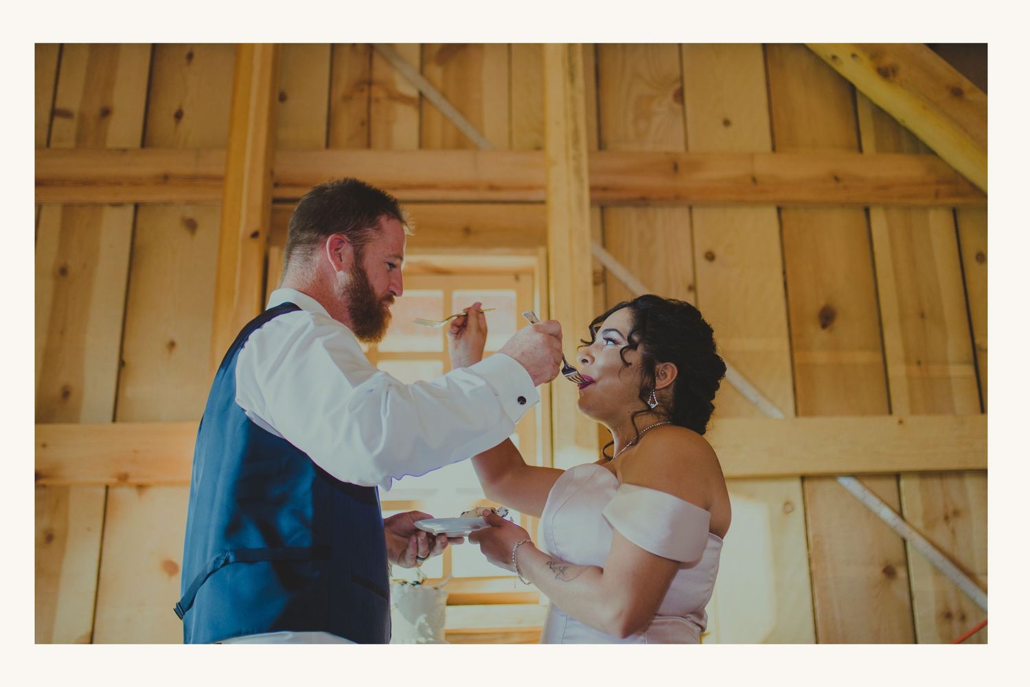 Bride and groom sharing a joyful cake tasting moment inside a rustic wooden barn at their wedding reception.