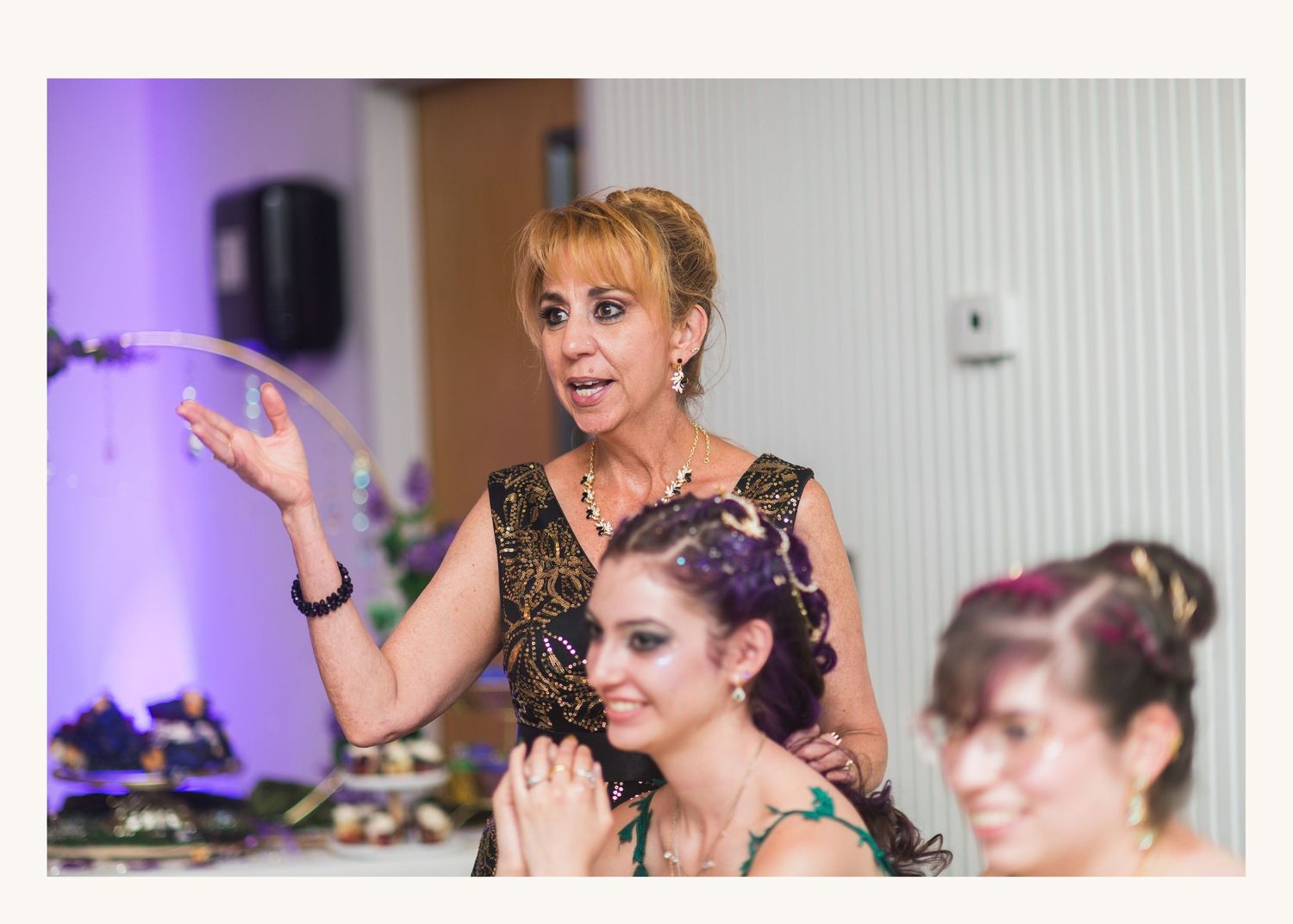 Woman in elegant black dress gesturing expressively at a formal event with guests seated nearby.