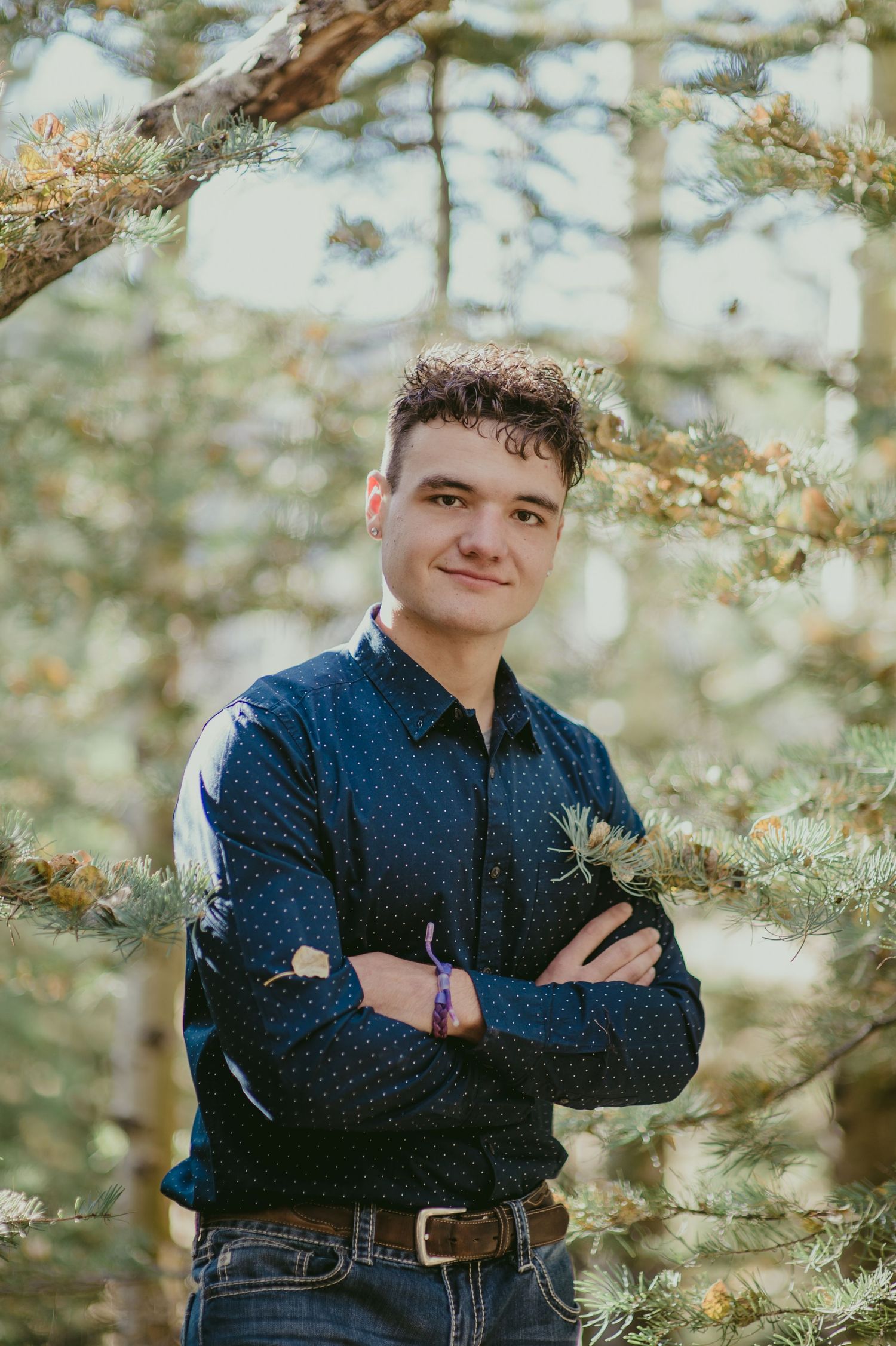 Young man in navy blue shirt stands with arms crossed among pine trees, smiling confidently outdoors.