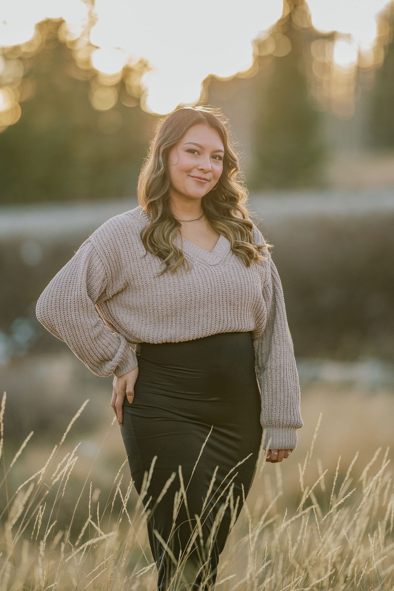 Person in a gray knit sweater and dark dress poses thoughtfully in golden sunlit field at sunset.