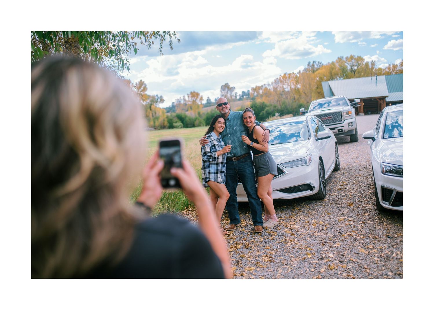 Someone taking a photo on their phone of people standing between parked cars on a gravel lot on an autumn day.