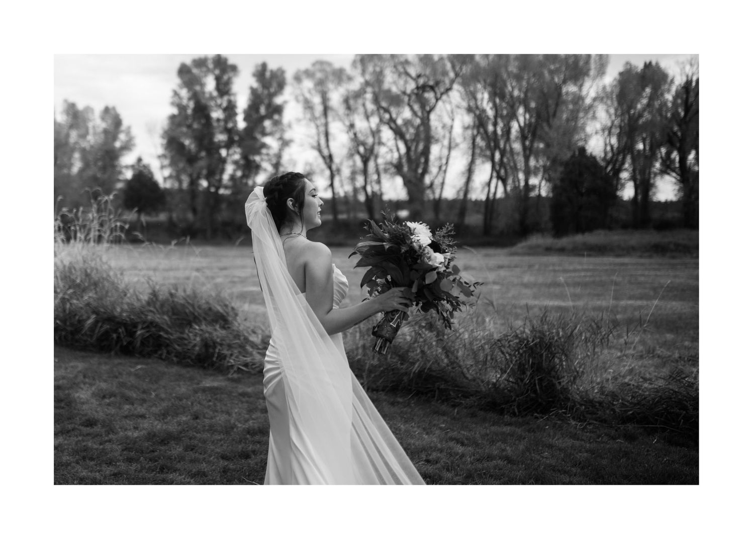 A black and white photo of a person in a flowing wedding dress holding a bouquet in a field at dusk.