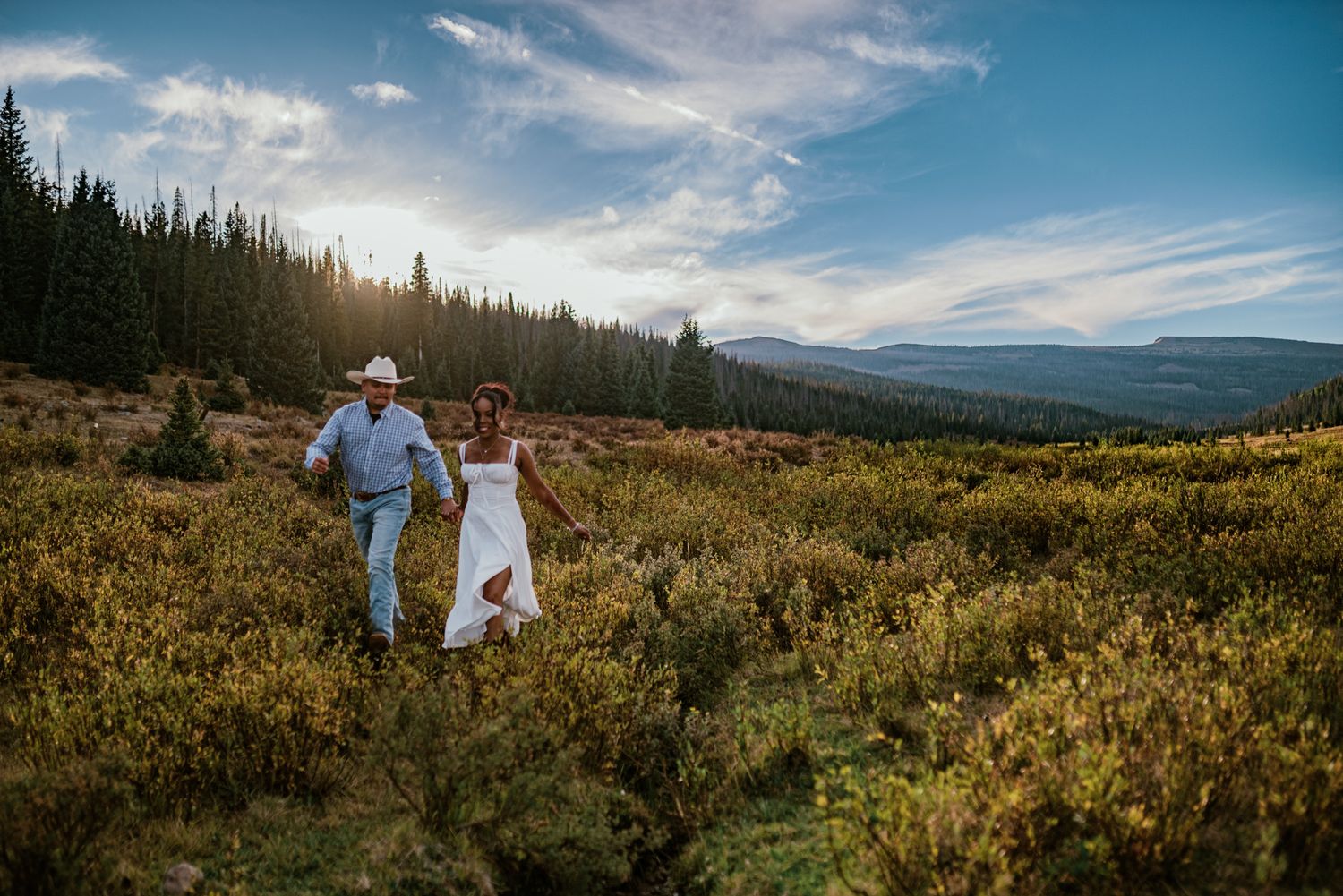 Couple in wedding attire walks hand-in-hand through mountain meadow at golden hour with pine forest backdrop.