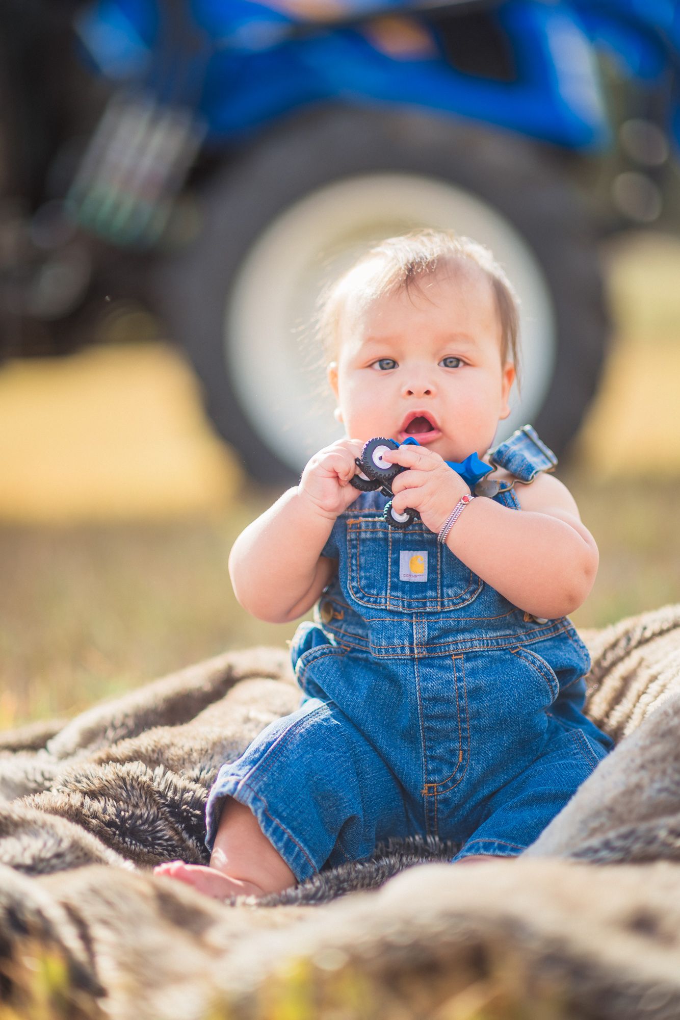 Adorable baby in denim overalls sitting on a blanket outdoors, chewing on a toy near a blue tractor.