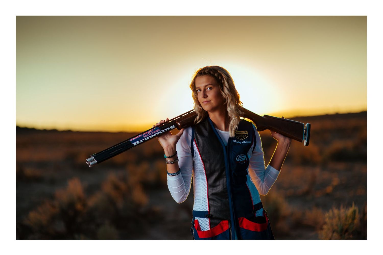 Woman holding a shotgun over her shoulder at sunset in a desert landscape, wearing a shooting vest.