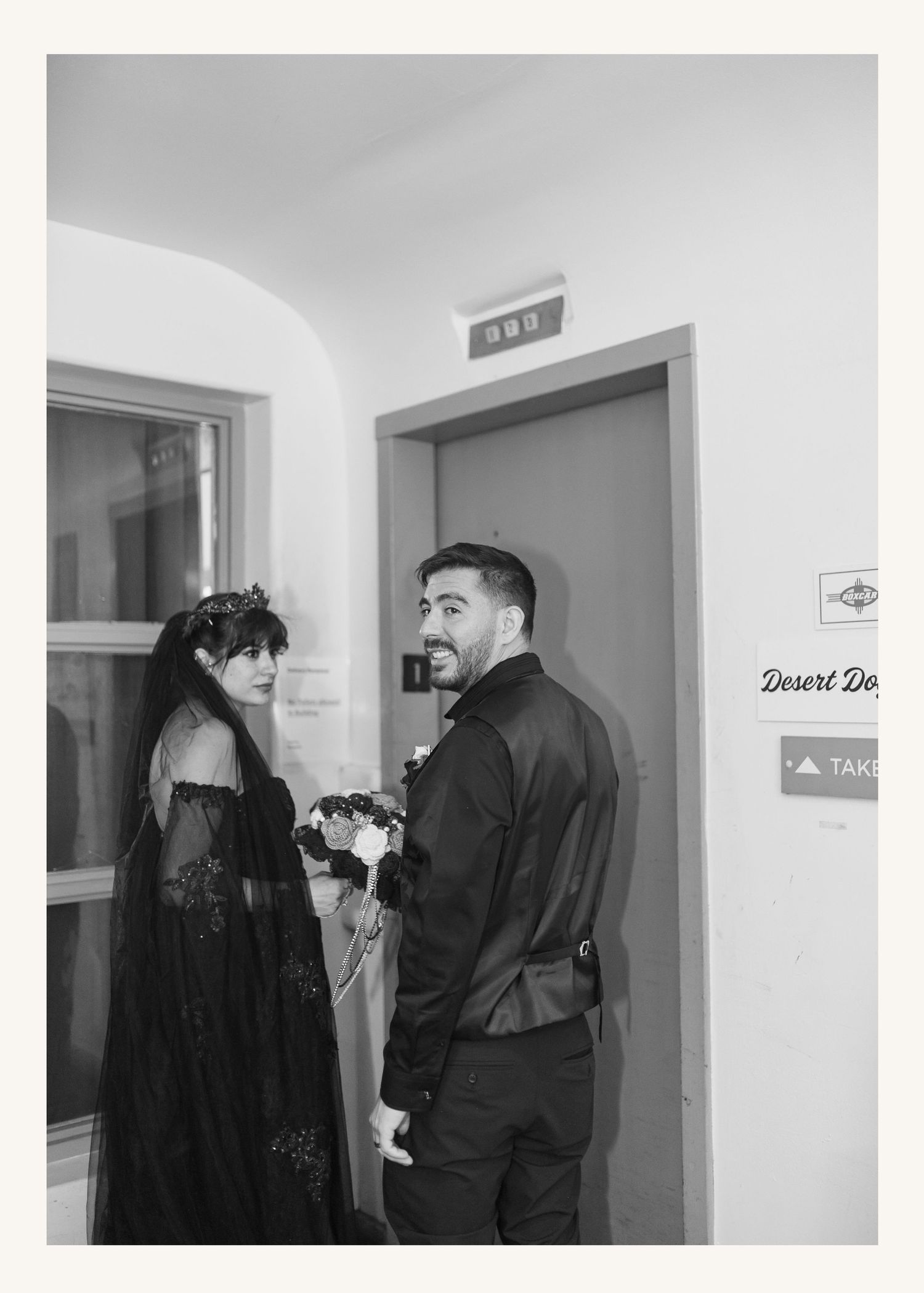 Black and white photo of a couple in formal attire, woman in black gown with floral headpiece holding bouquet near elevator.