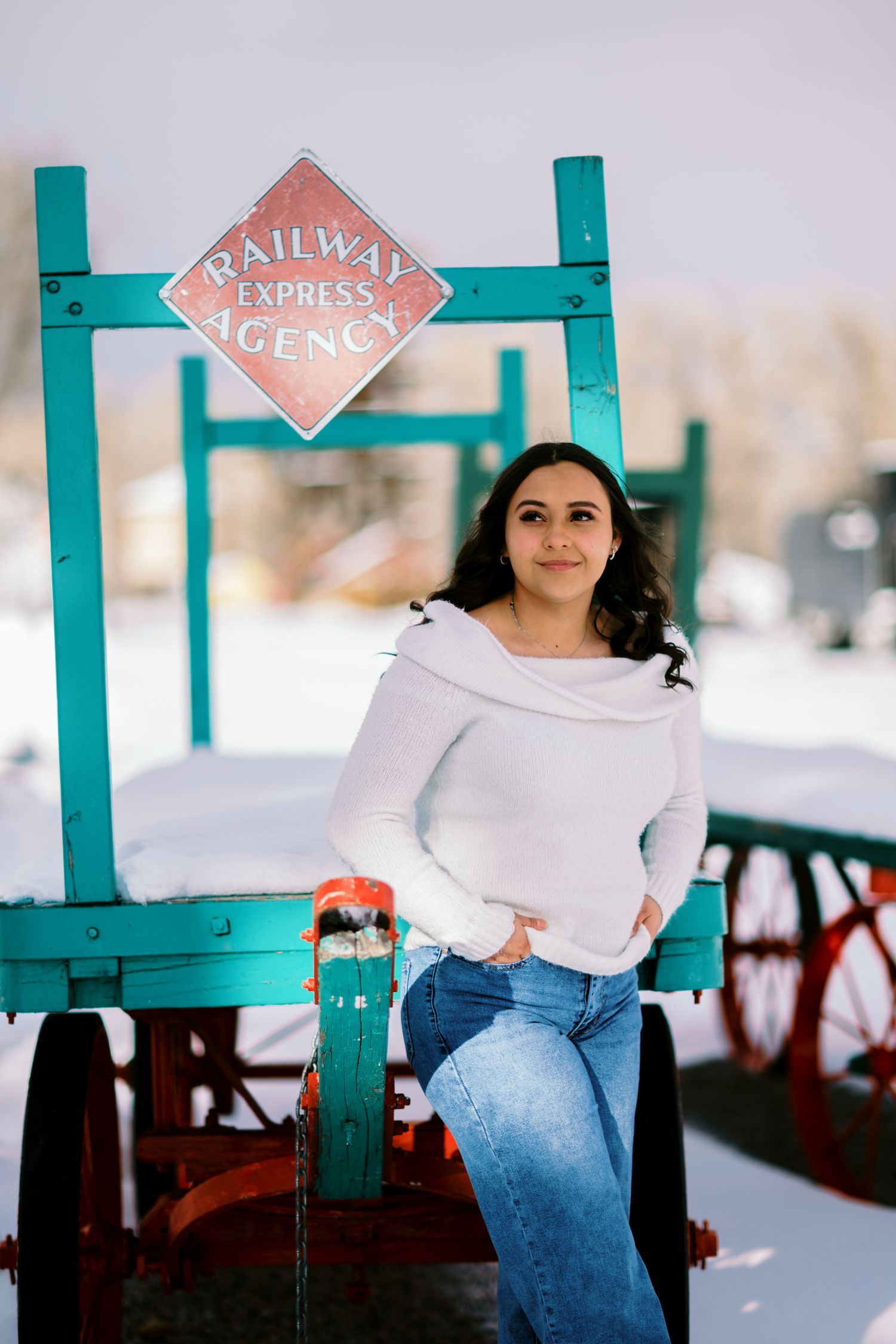 A person in a white sweater and jeans poses against a turquoise-framed structure in a snowy winter setting.