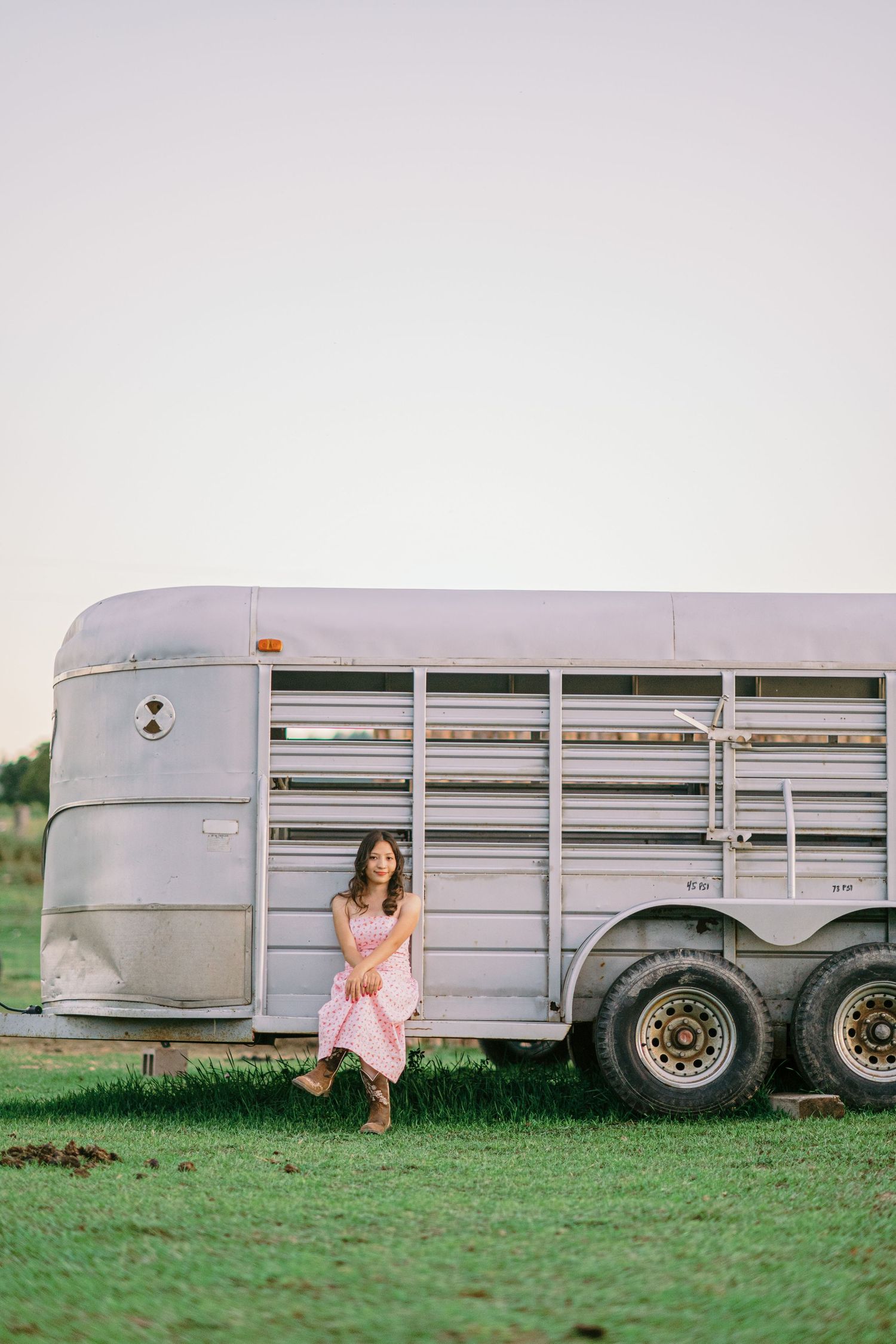 Four sequential photos show someone in a pink dress playing near a silver livestock trailer in a grassy field.