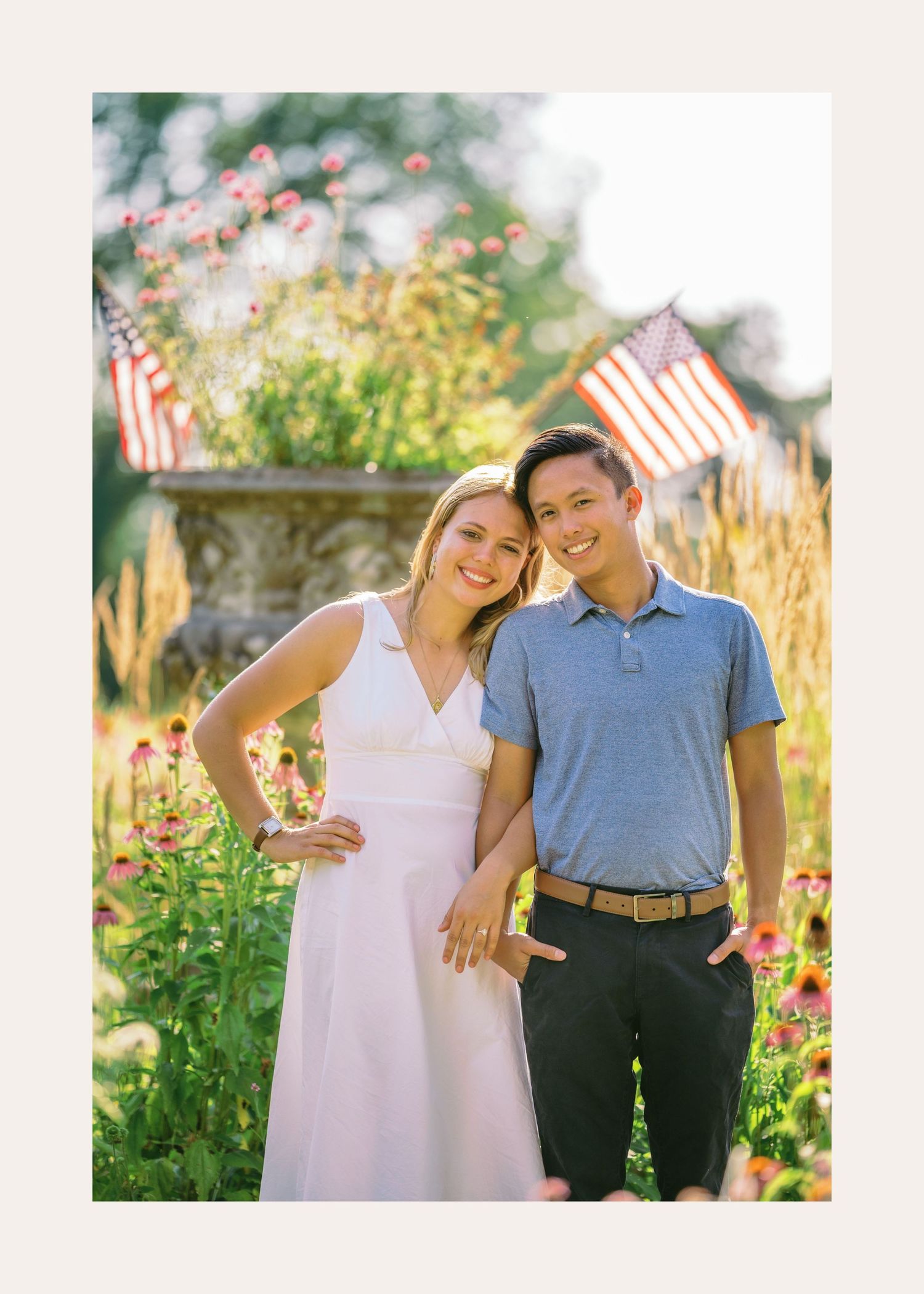 A couple in summer attire poses together in a garden with small American flags and flowers in the background.