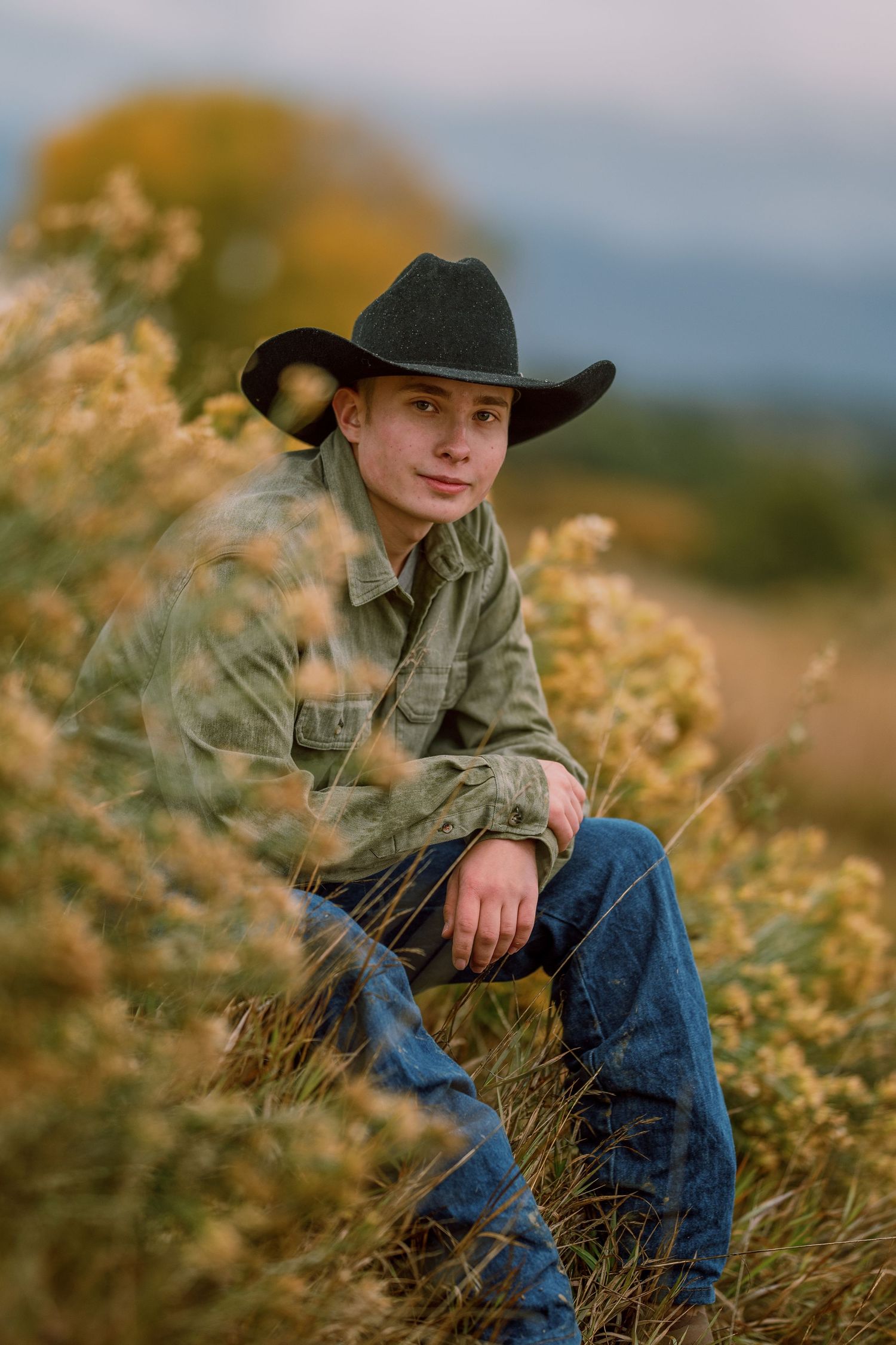 A person in western attire with cowboy hat kneels among tall grasses during golden hour in a rustic outdoor setting.