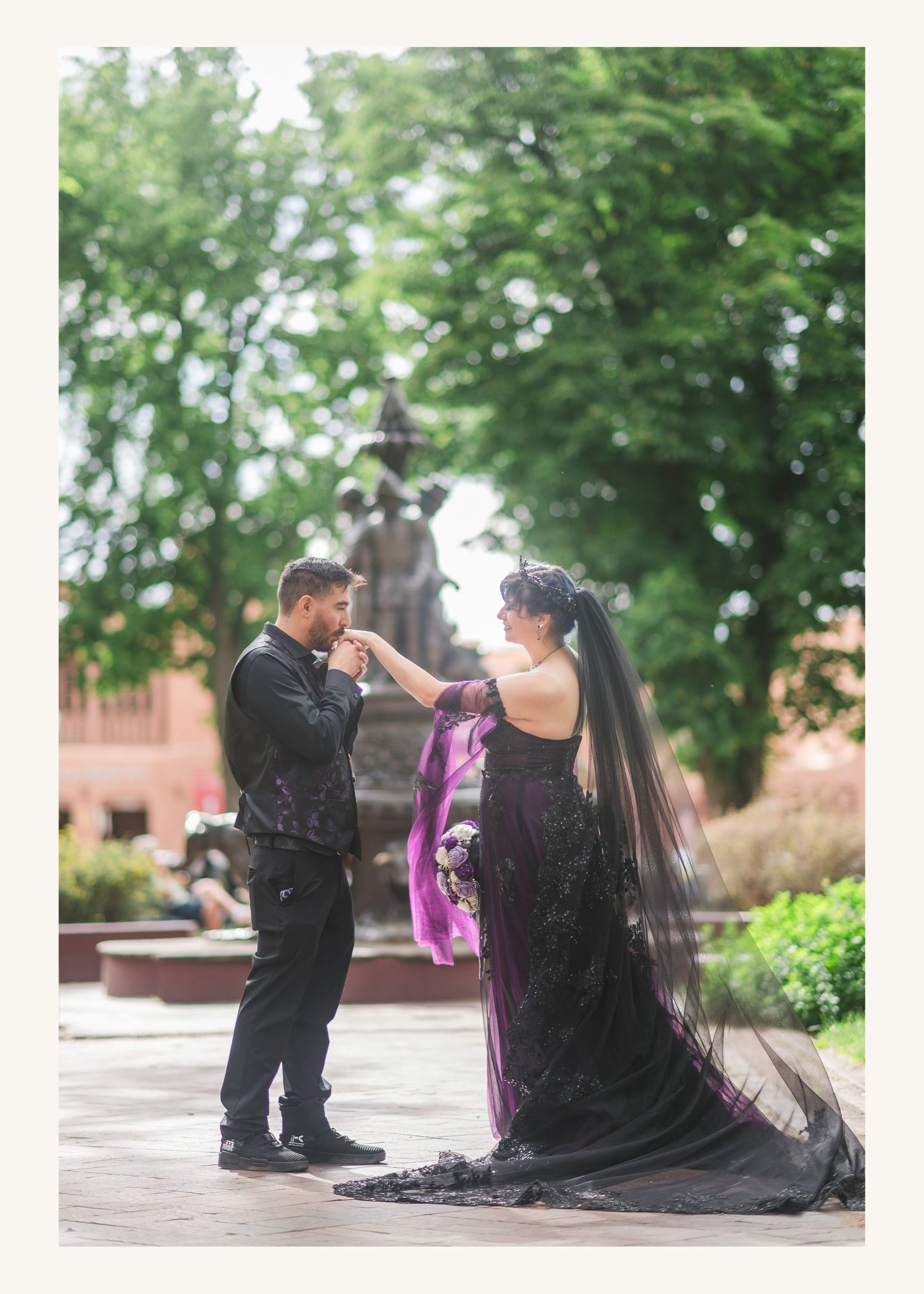 Gothic wedding couple in black and purple attire sharing a romantic moment by a fountain in a lush garden.