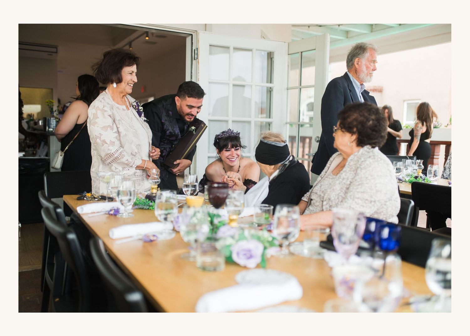 Guests socializing at an elegant dinner party, gathered around a long table set with glassware in a bright venue.