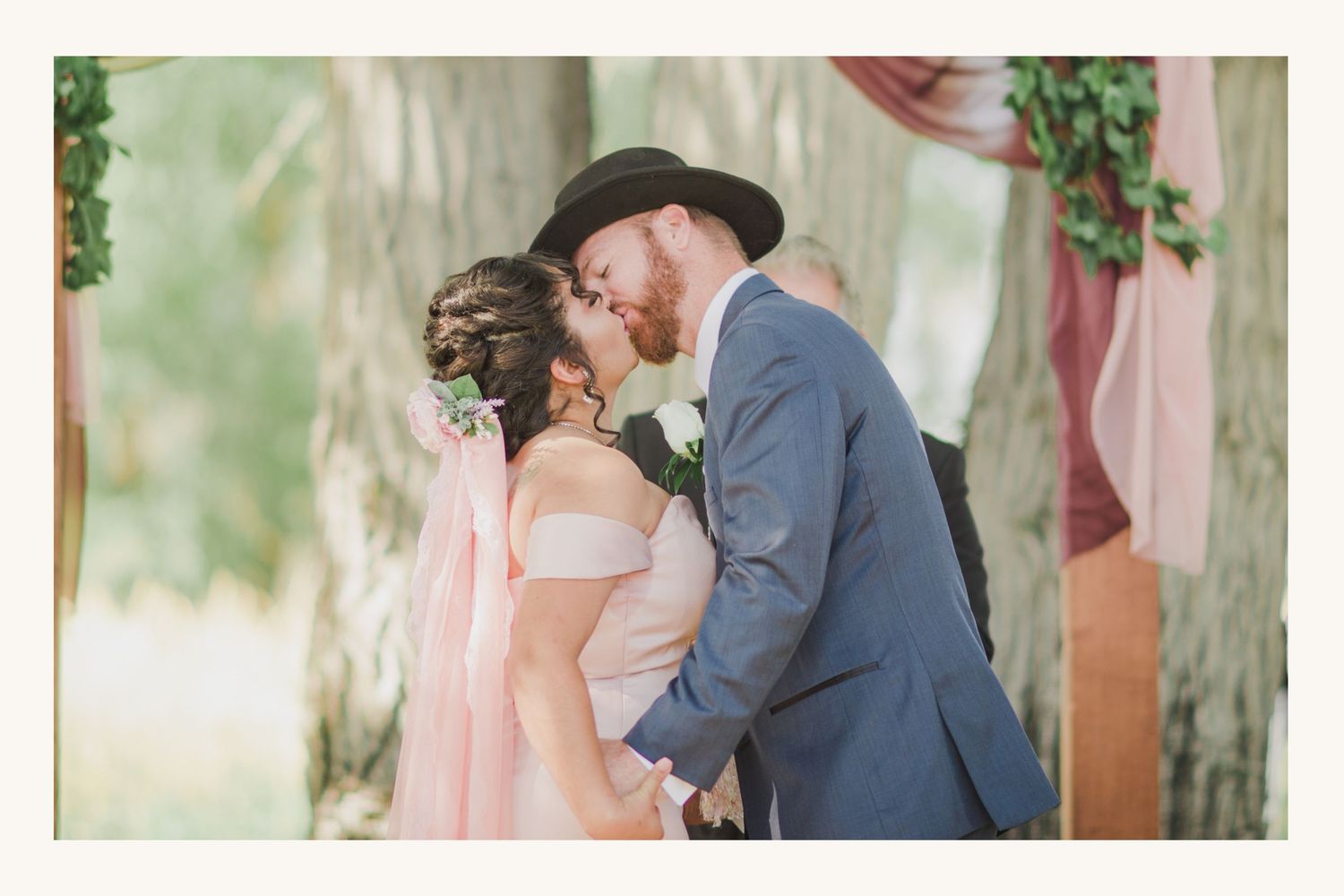 Bride and groom share first kiss at outdoor wedding ceremony beneath tree with dusty rose ribbon decor.