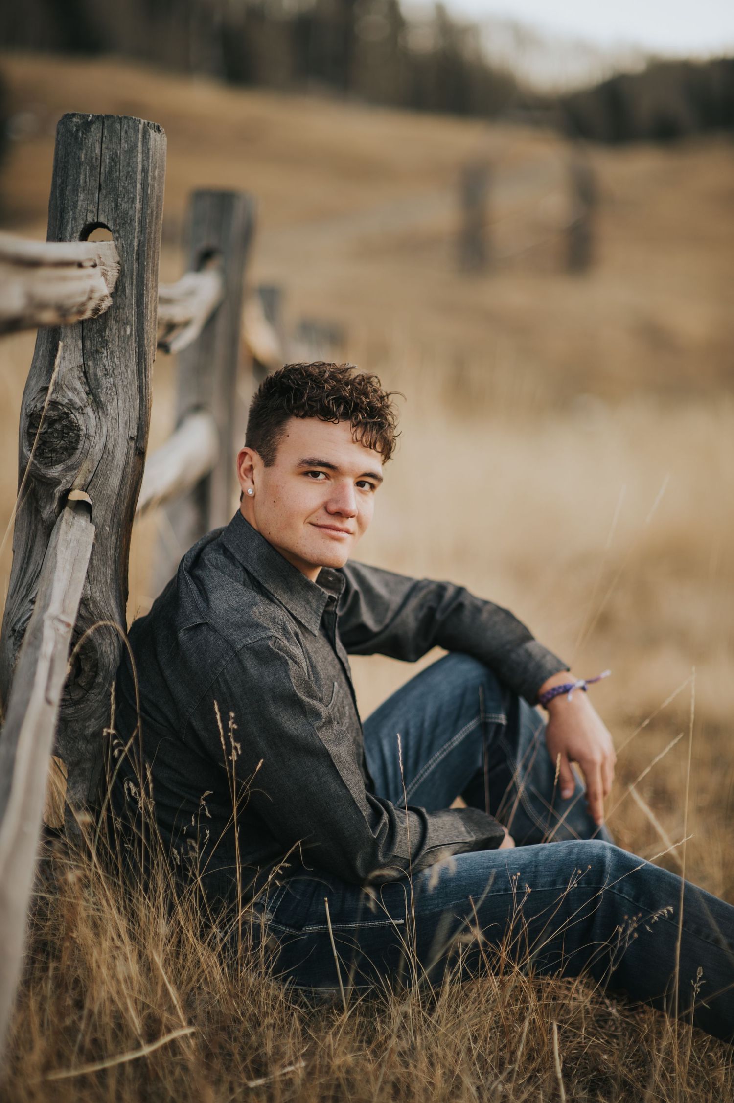 Young man in gray shirt and jeans sitting by a rustic wooden fence in a golden field.