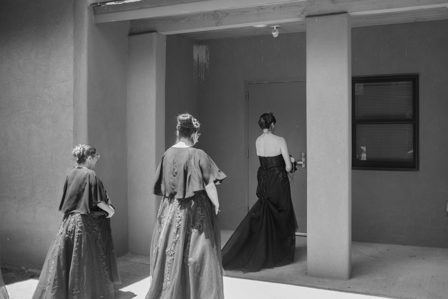 Three women in elegant period gowns stand near a doorway in a black and white architectural setting.