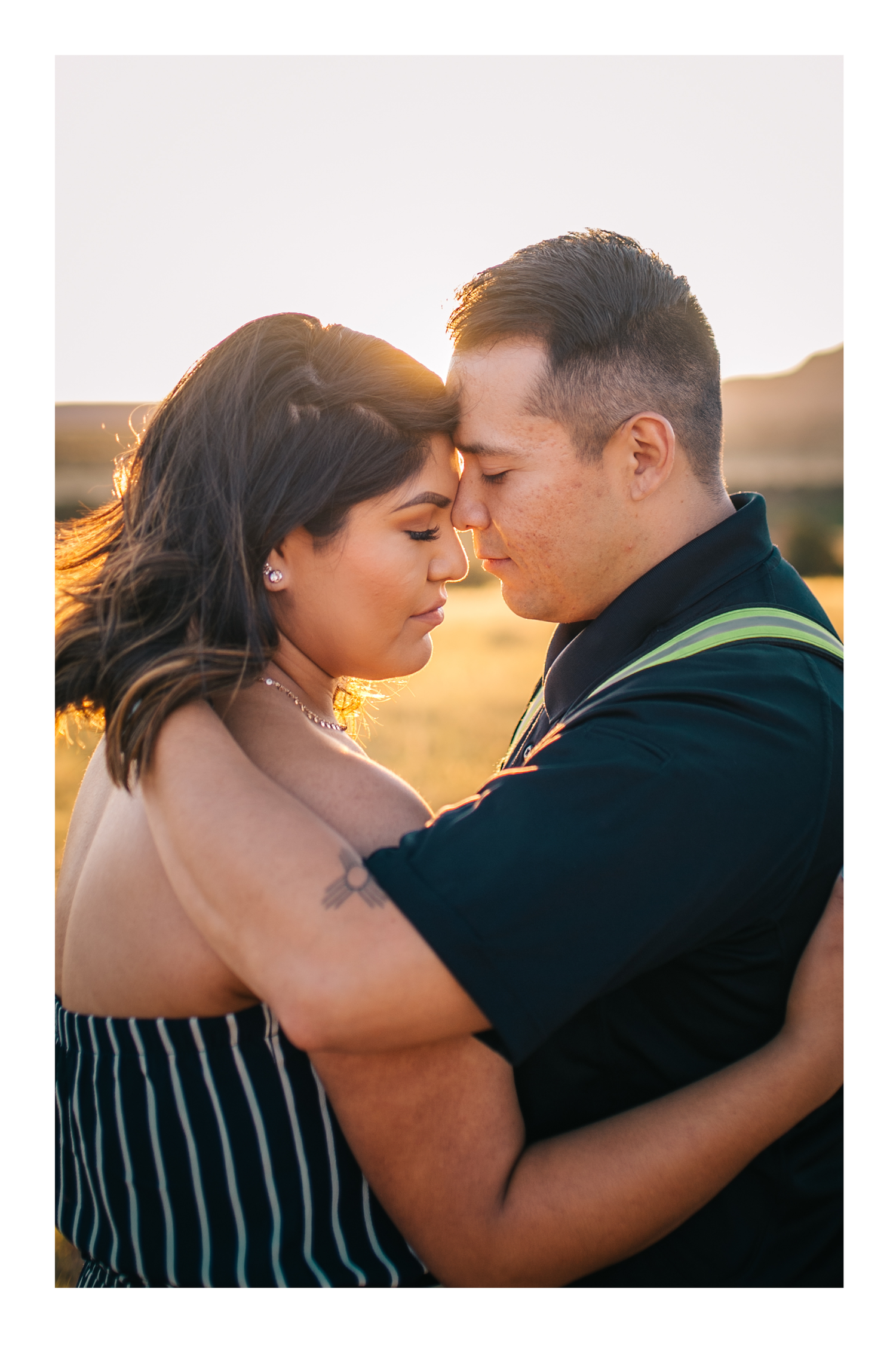 Couple embracing at golden hour in a field, foreheads touching romantically, woman with tattoo in striped top.