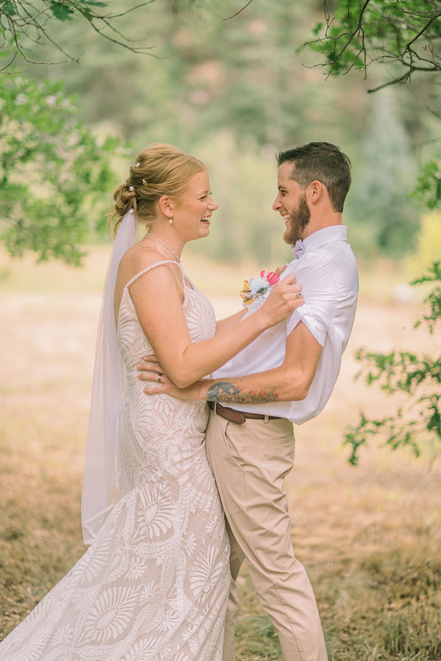 A smiling bride in a lace gown and veil embraces a bearded groom in a white shirt outdoors.