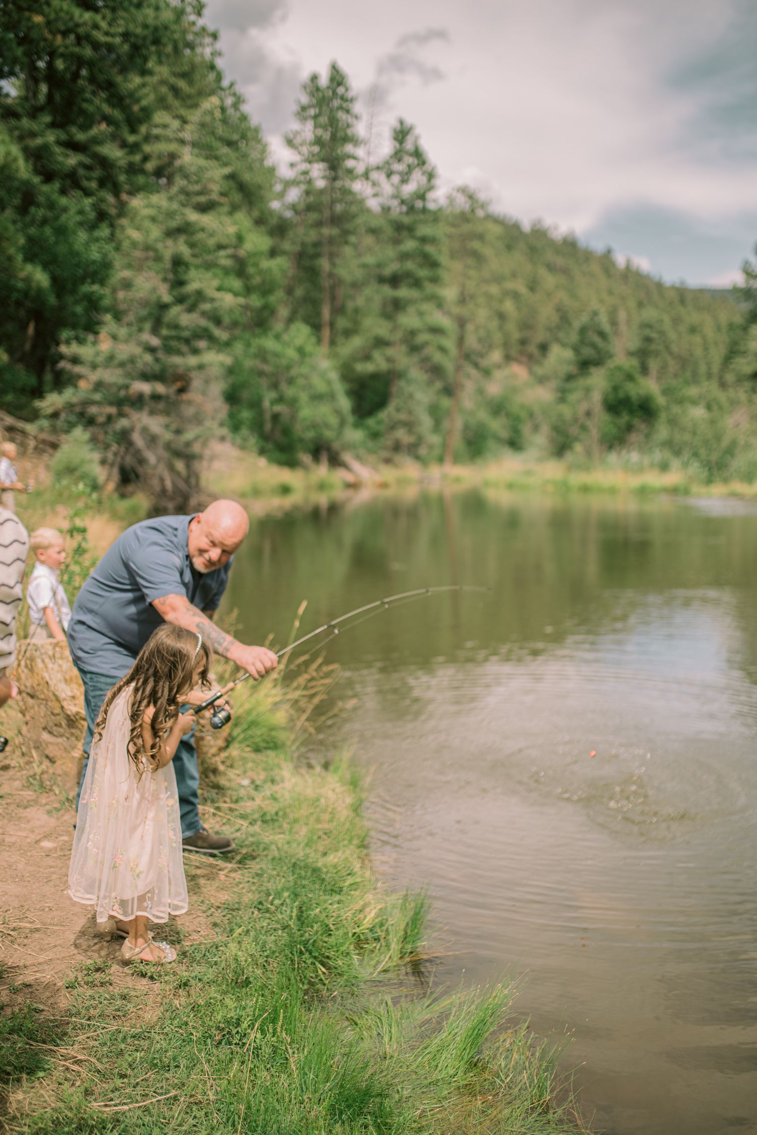 A man teaches a young girl to fish by a serene mountain river surrounded by lush green forest.