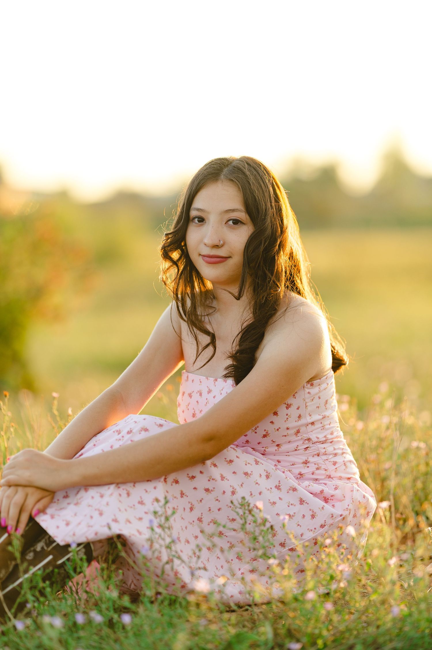 A person in a floral pink dress sits gracefully in a sunlit meadow among wildflowers at golden hour.