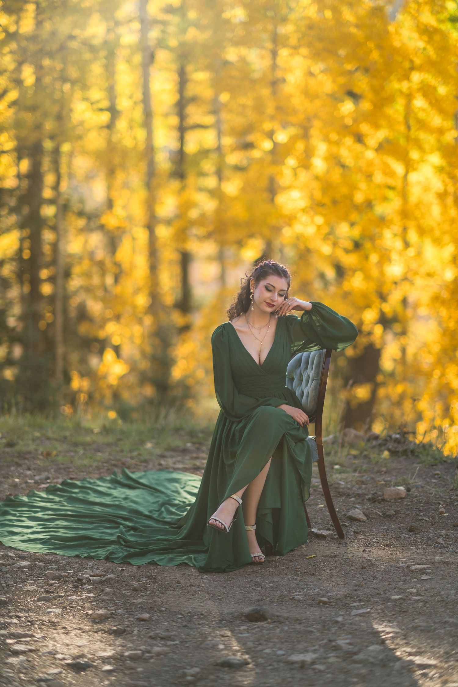 A woman in an emerald green dress poses gracefully on forest ground surrounded by golden autumn foliage.