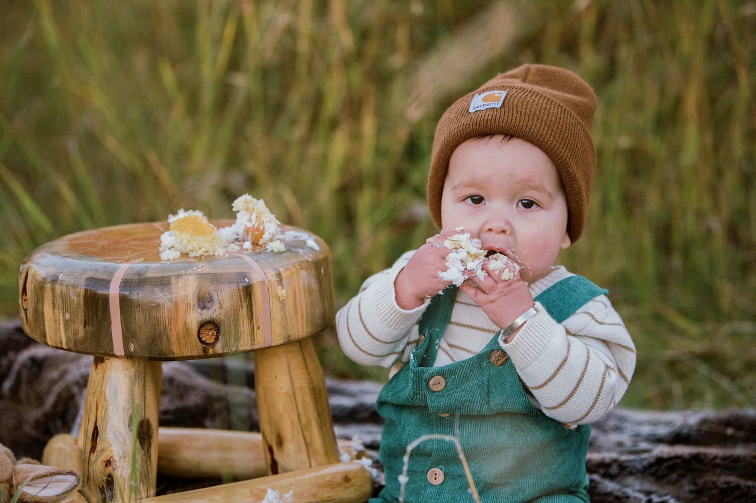 Figure in brown beanie and green overalls posed near wooden outdoor setting.