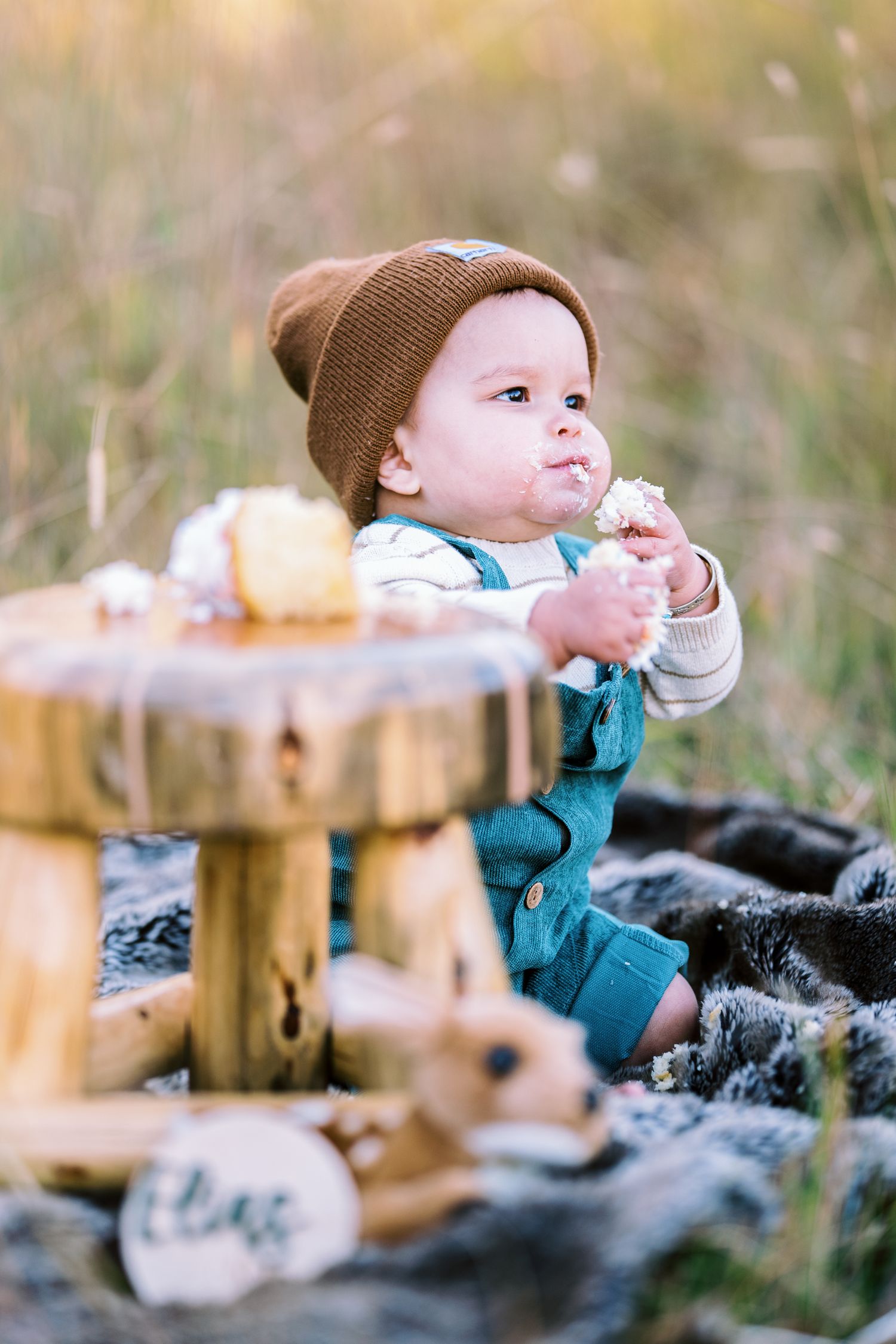 A baby in teal overalls and brown beanie sits by a wooden stool in a rustic outdoor setting.