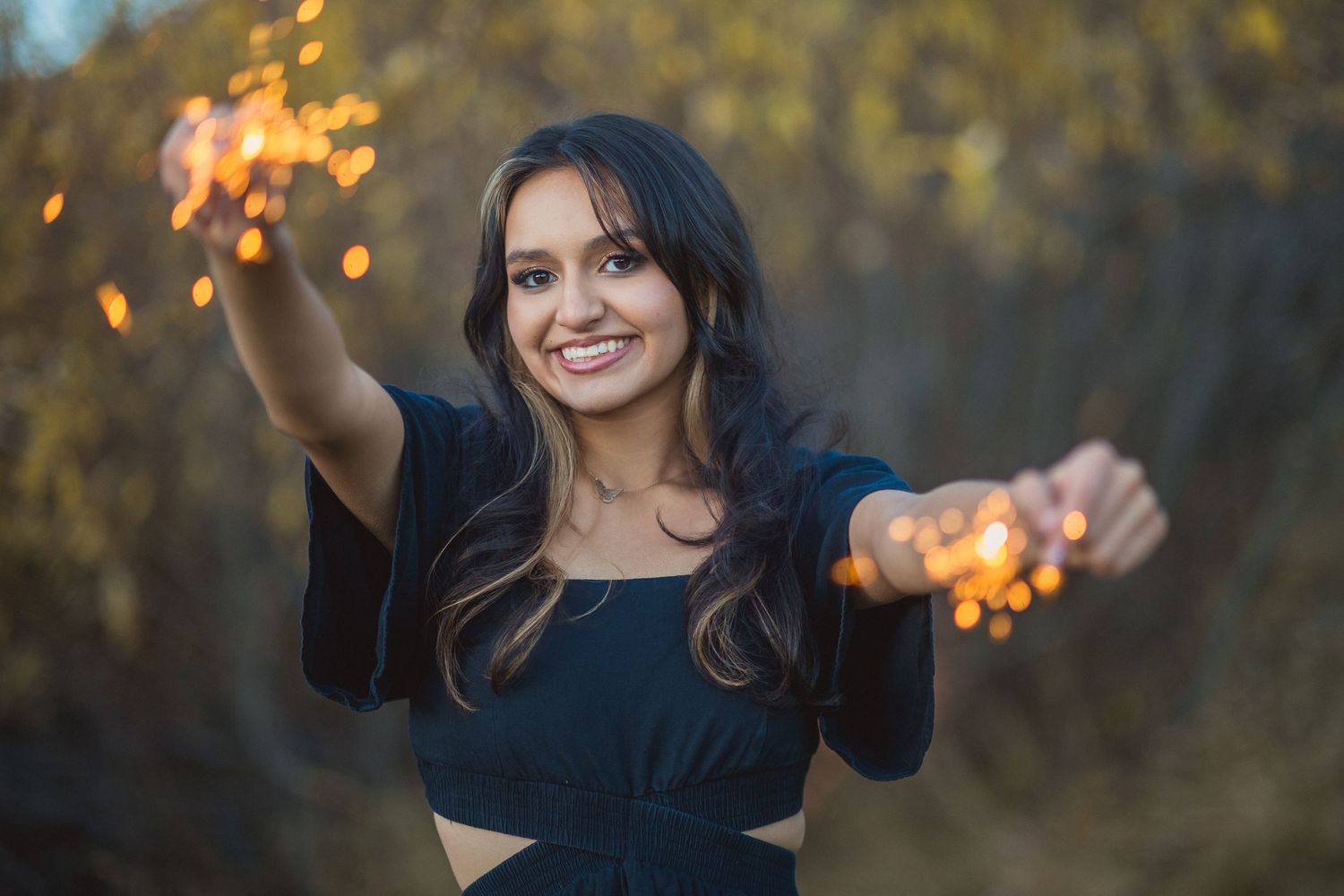 Person in black outfit holds sparklers with outstretched arms during golden hour outdoors.