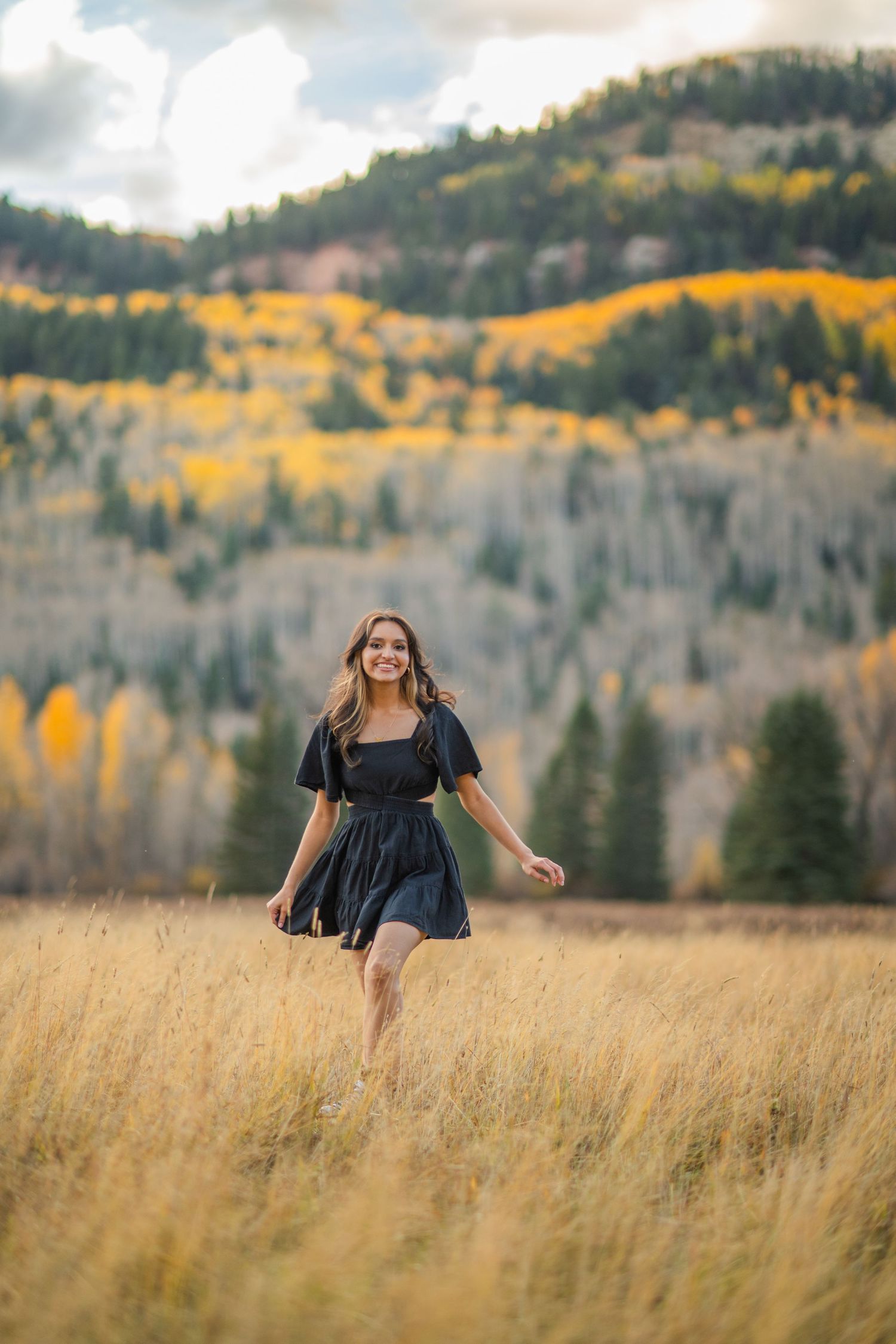 A person in a dark dress spins joyfully through a golden autumn field with mountains and fall foliage in the background.
