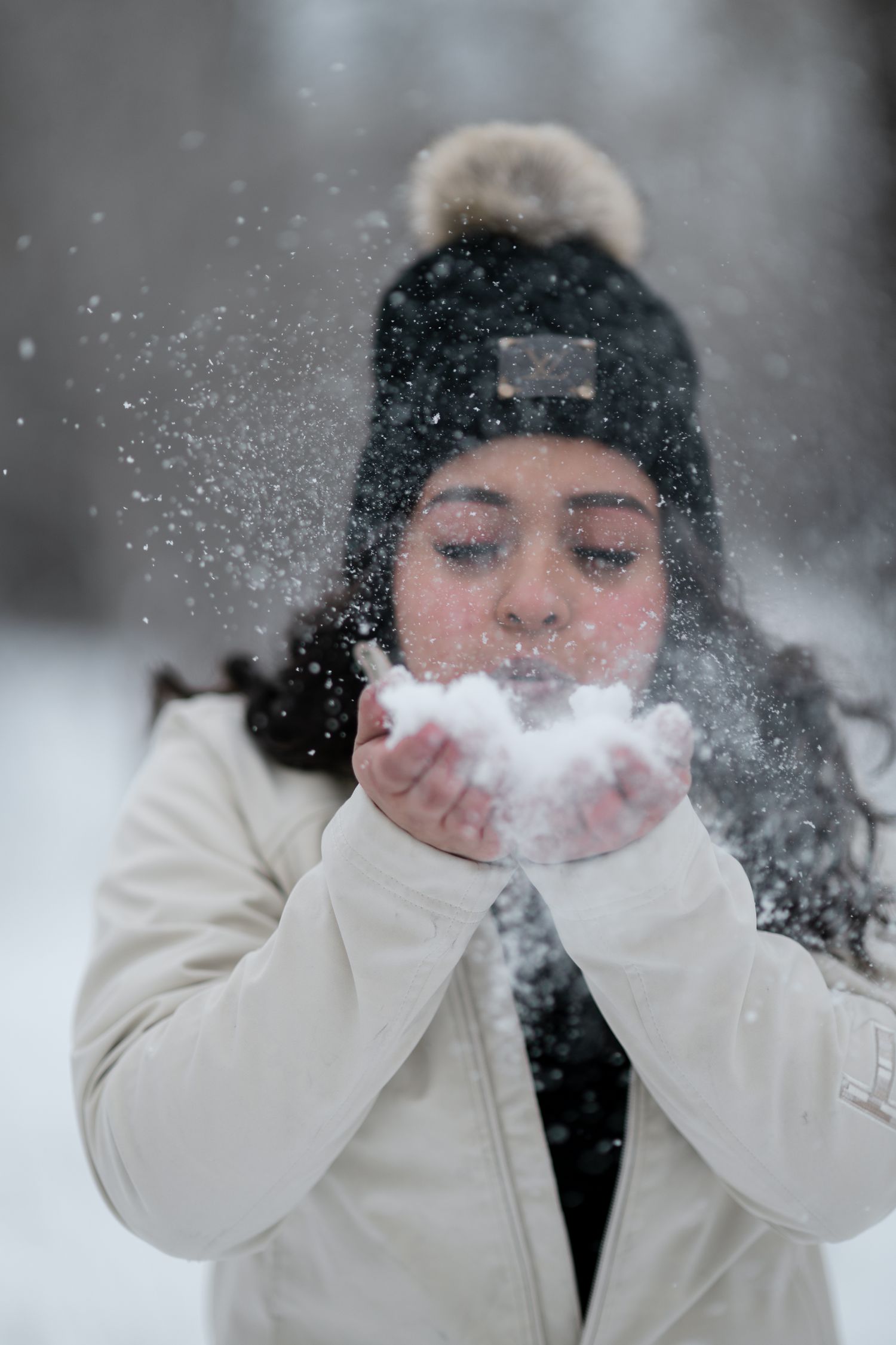 Someone in a white winter coat blows snow from their hands while snowflakes fall around them in a magical winter scene.