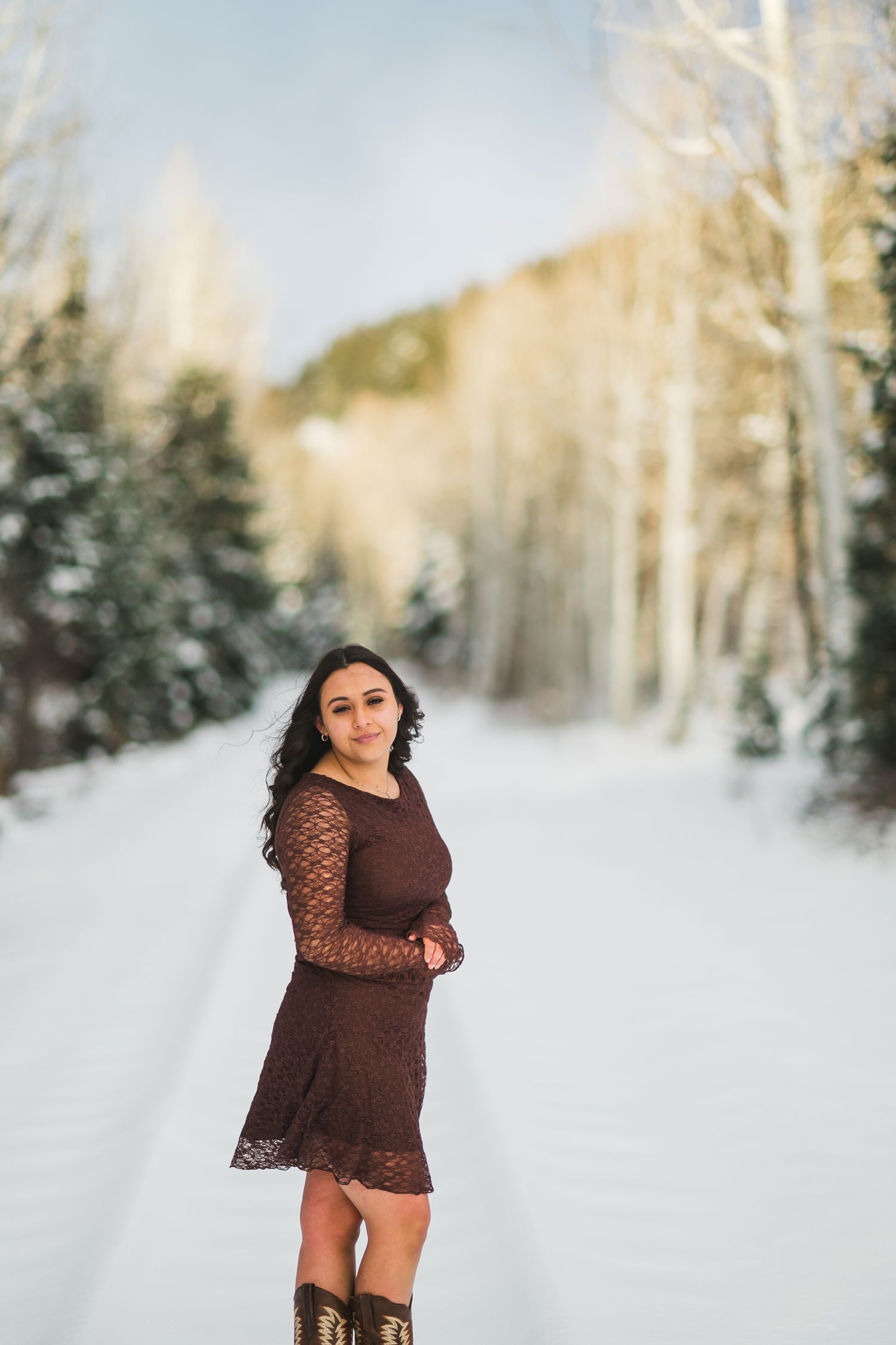 A woman in a brown lace dress poses in a snowy winter forest setting with sunlit trees in the background.