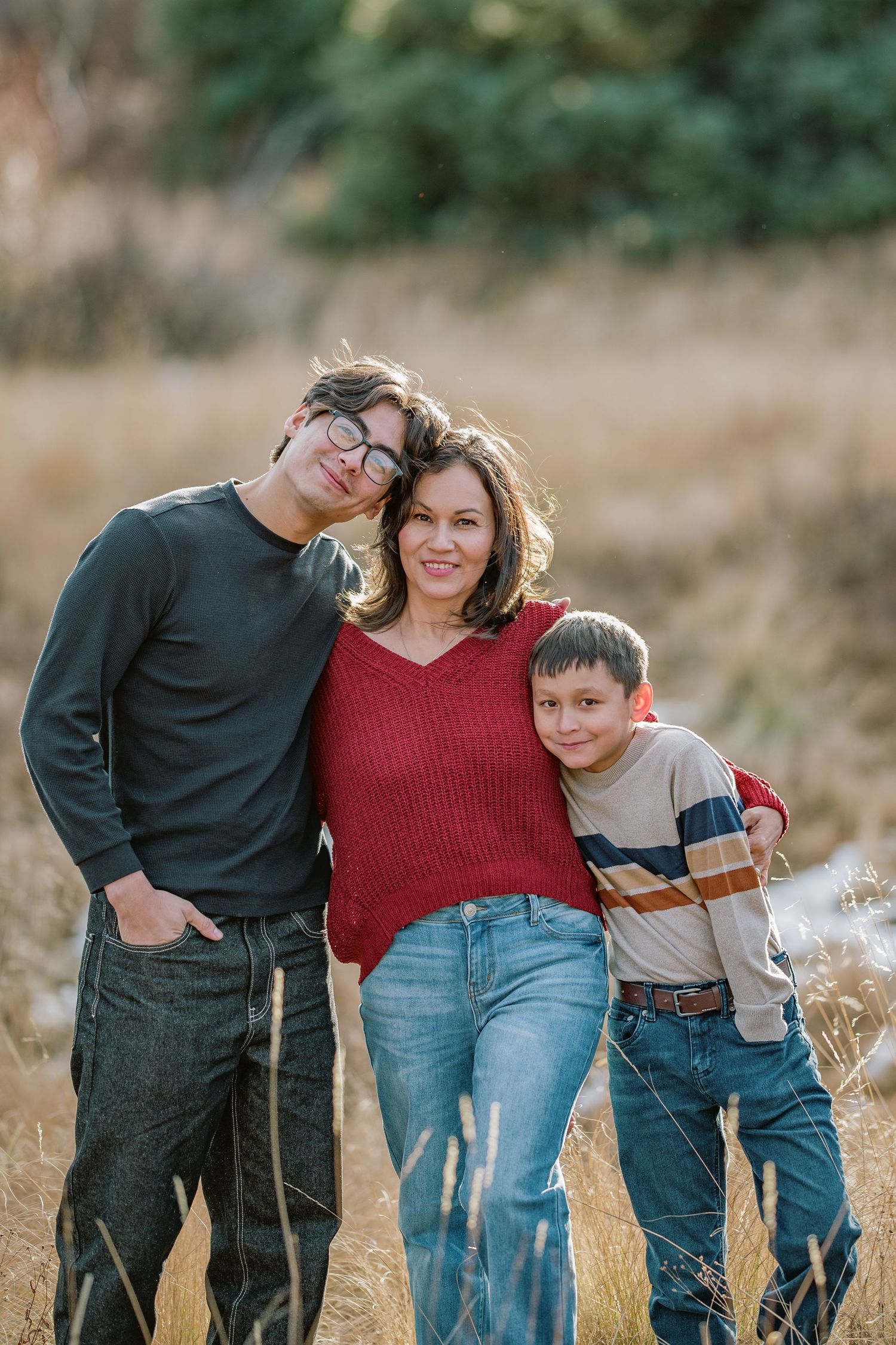 A family of three poses together in a field wearing casual clothing on an autumn day.