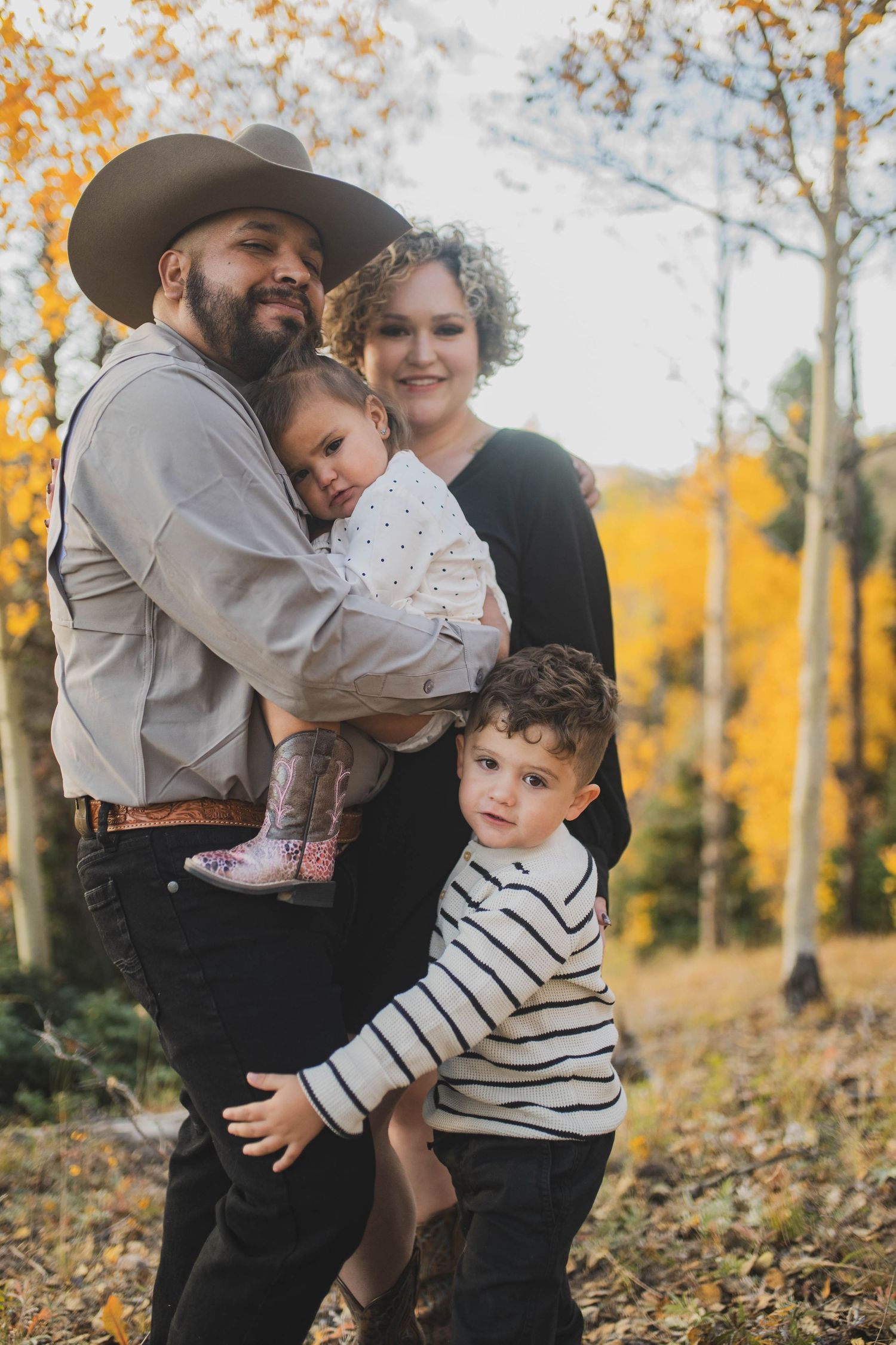 A family poses together against a backdrop of autumn aspen trees with golden yellow leaves falling in a forest setting.