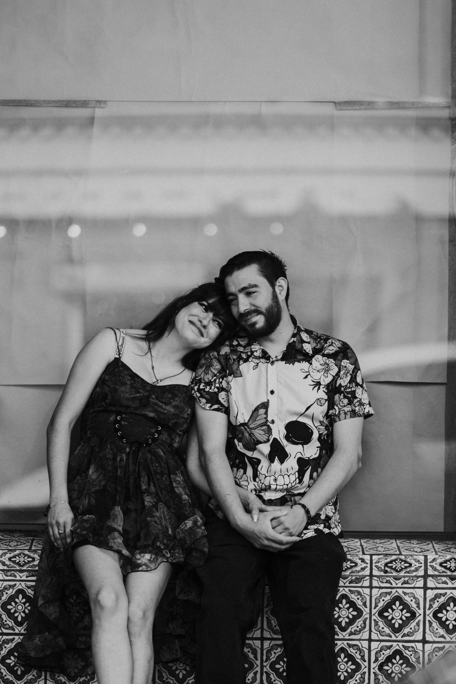 Black and white photo of a couple sitting together, woman resting head on bearded man's shoulder, tiled floor background.