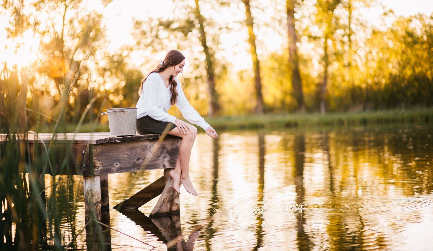 A figure sits on a wooden dock by a serene lake at sunset wearing white and dark clothing.