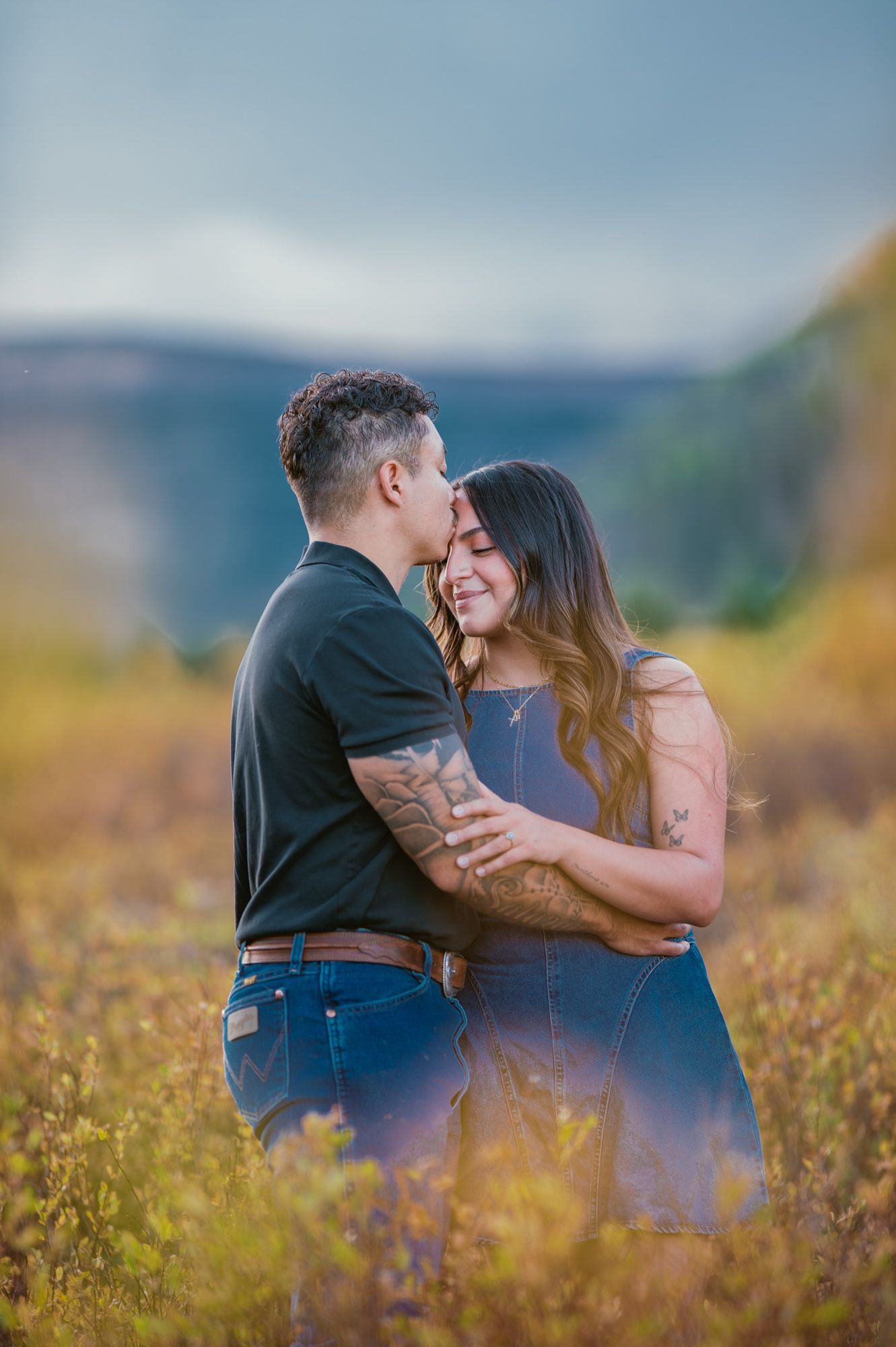 Couple embracing in golden autumn field, man kissing woman's forehead, mountains in background.