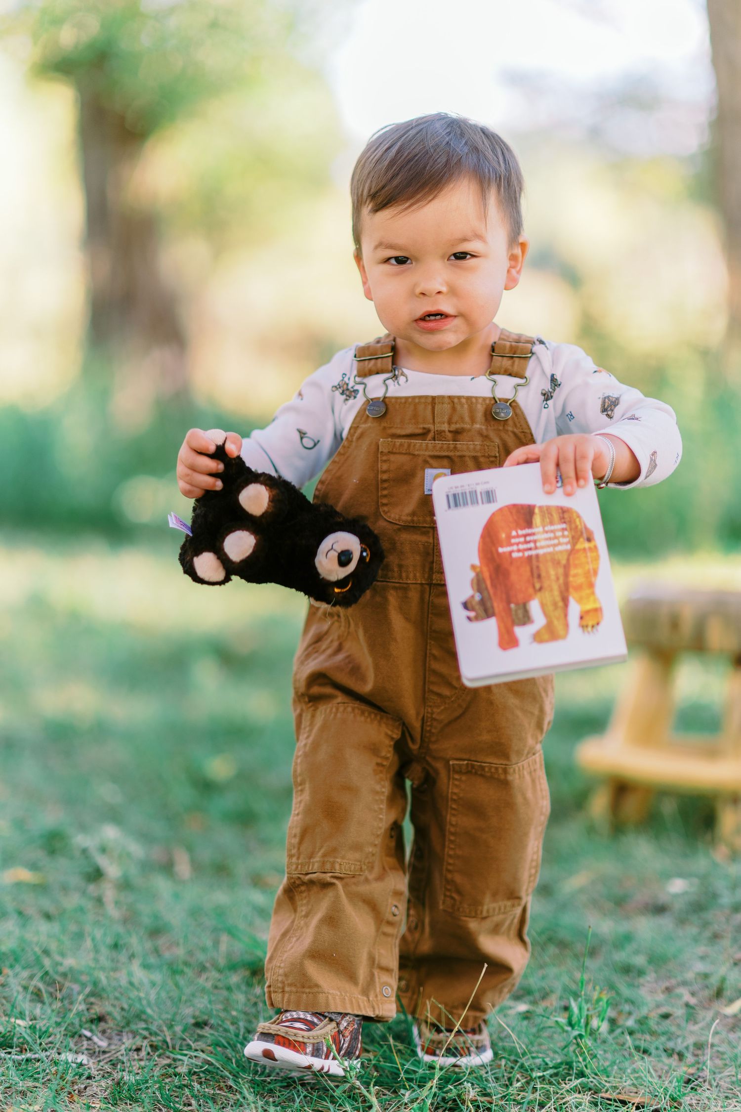 Small child wearing brown overalls and holding a plush toy stands on grass in an outdoor setting.