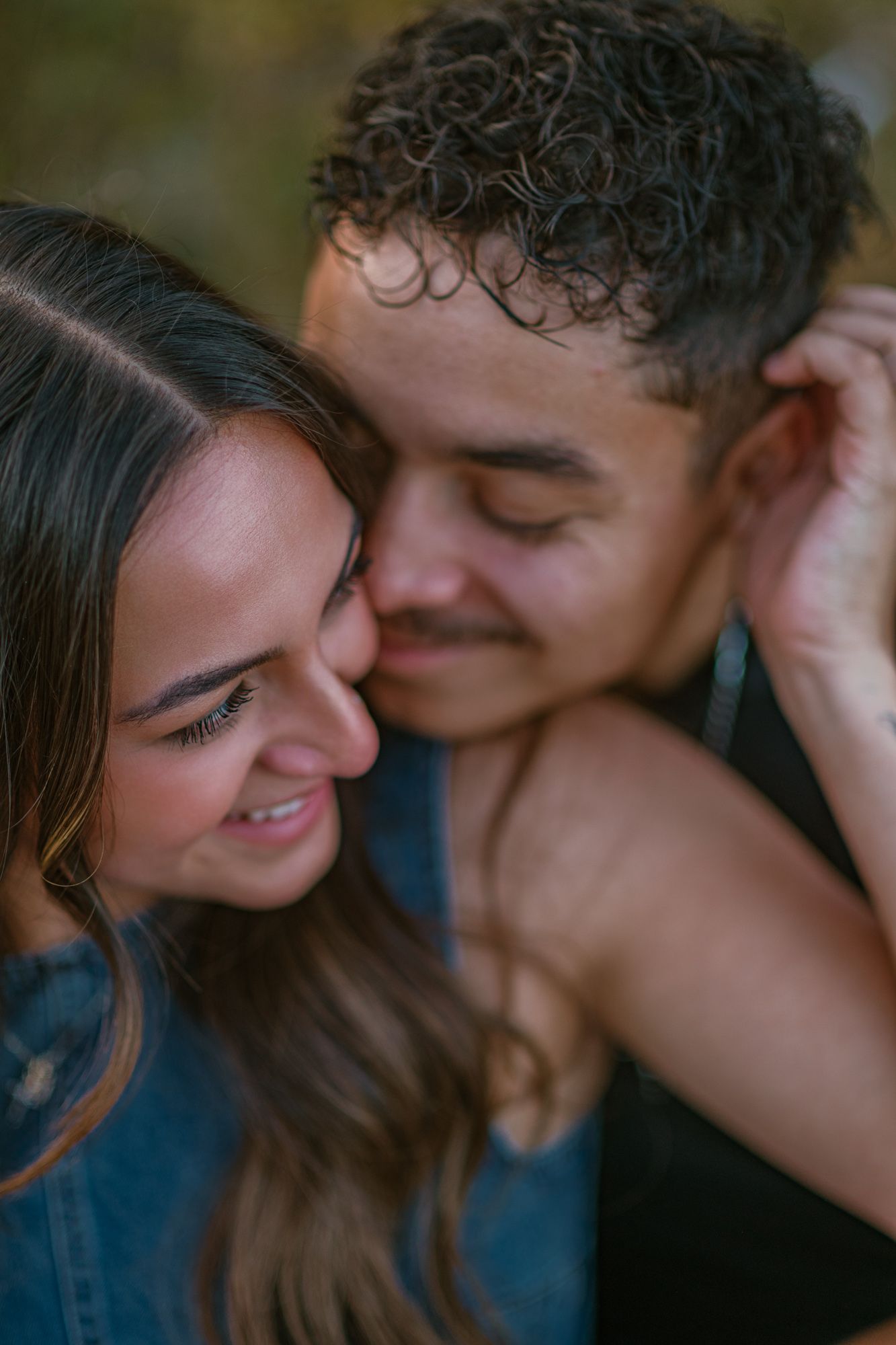 A couple shares an intimate moment, the man nuzzling the smiling woman's cheek outdoors.