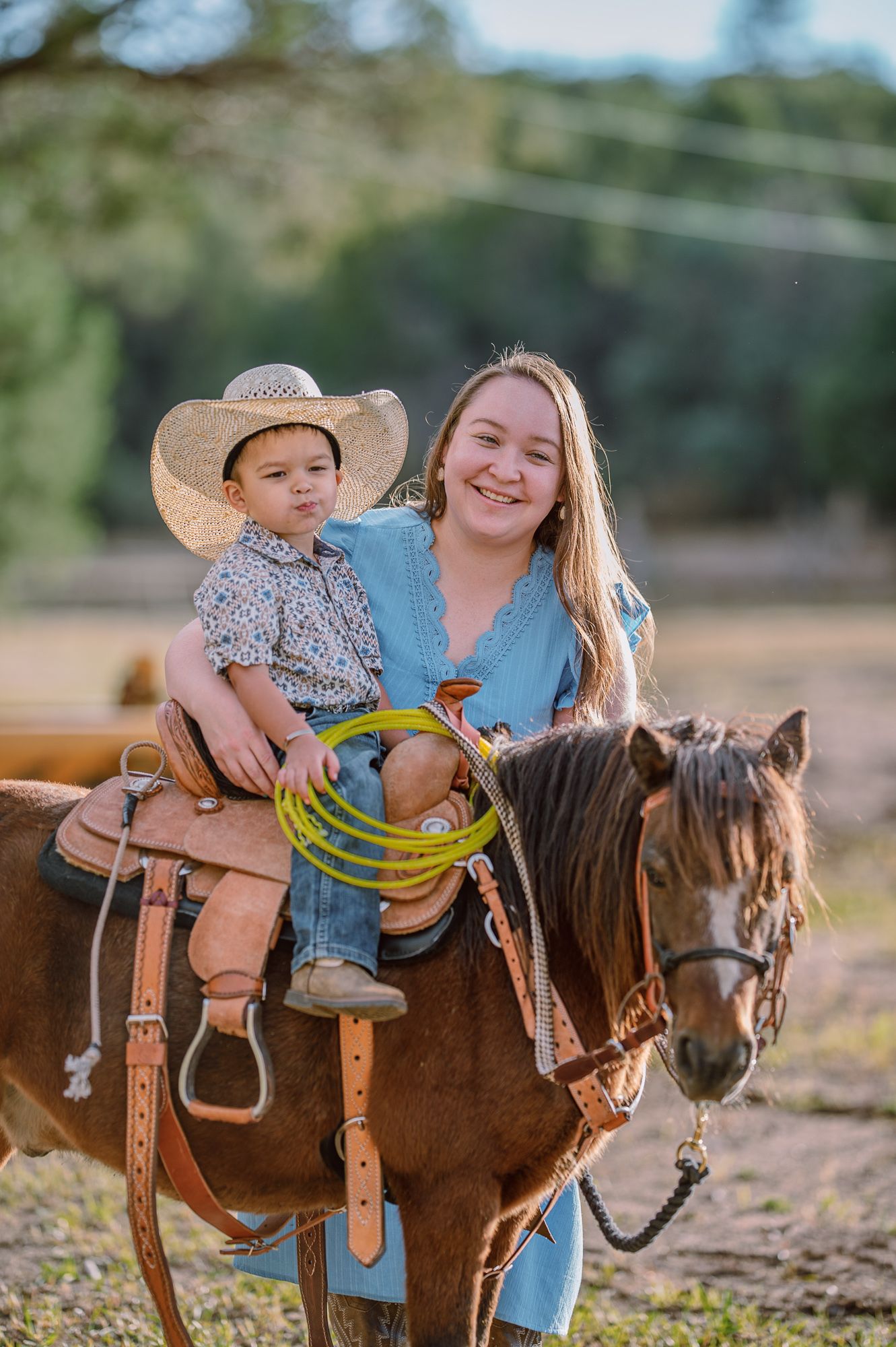 Young woman and toddler in cowboy hat sitting on brown horse with lasso, smiling outdoors on sunny day.