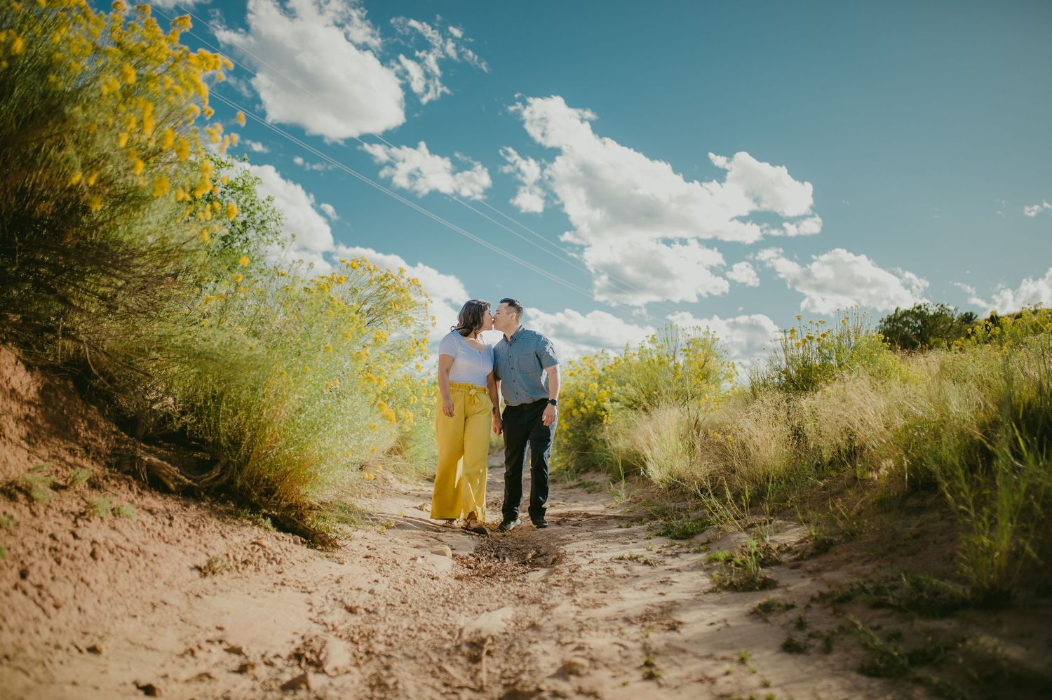 Couple embracing on a desert trail surrounded by yellow flowers and blue sky.