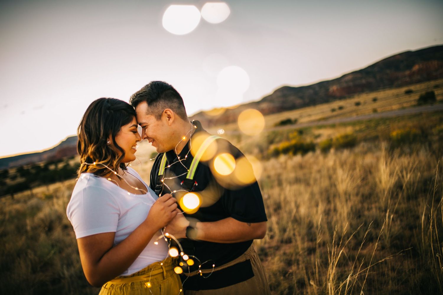 Couple sharing intimate moment at sunset with lens flare and mountain backdrop.