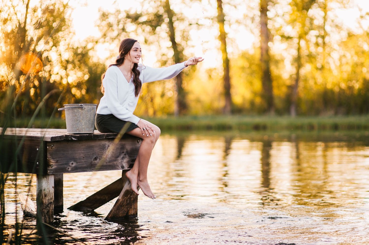 Person sitting on wooden dock points across calm lake waters during golden sunset.