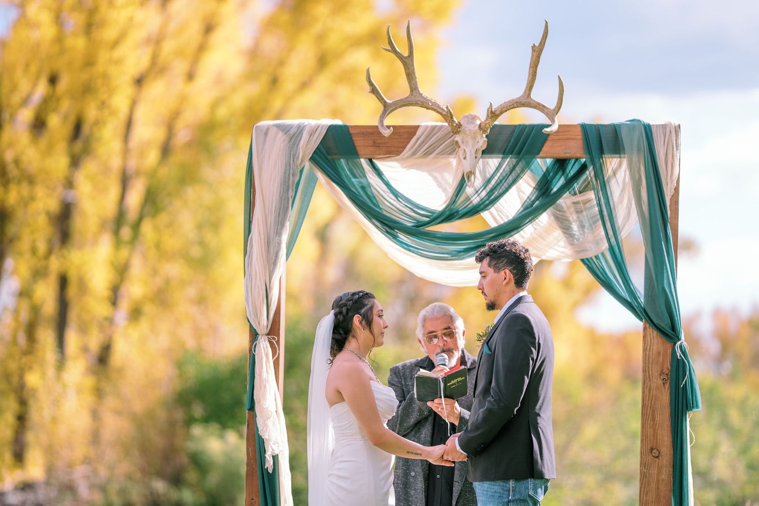 Wedding ceremony under an arch decorated with antlers and teal fabric against autumn trees.