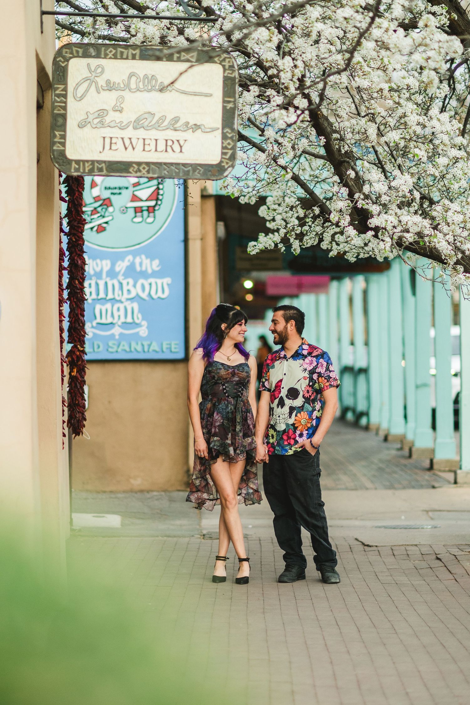 Couple holding hands on a Santa Fe sidewalk under blooming white cherry blossoms near a colorful jewelry shop.