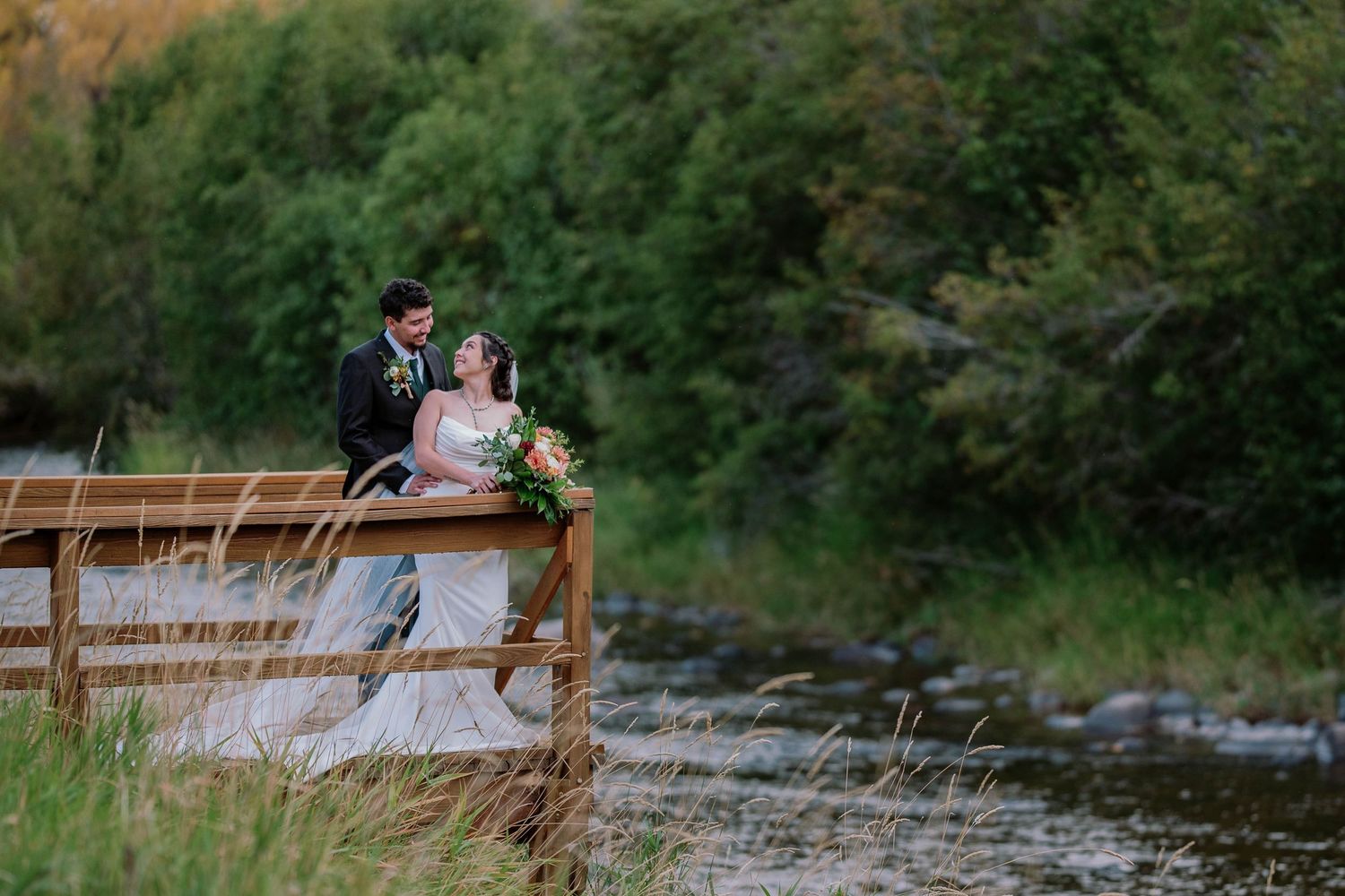 Two people in formal wedding attire sit together on a wooden dock near a stream at sunset surrounded by greenery.