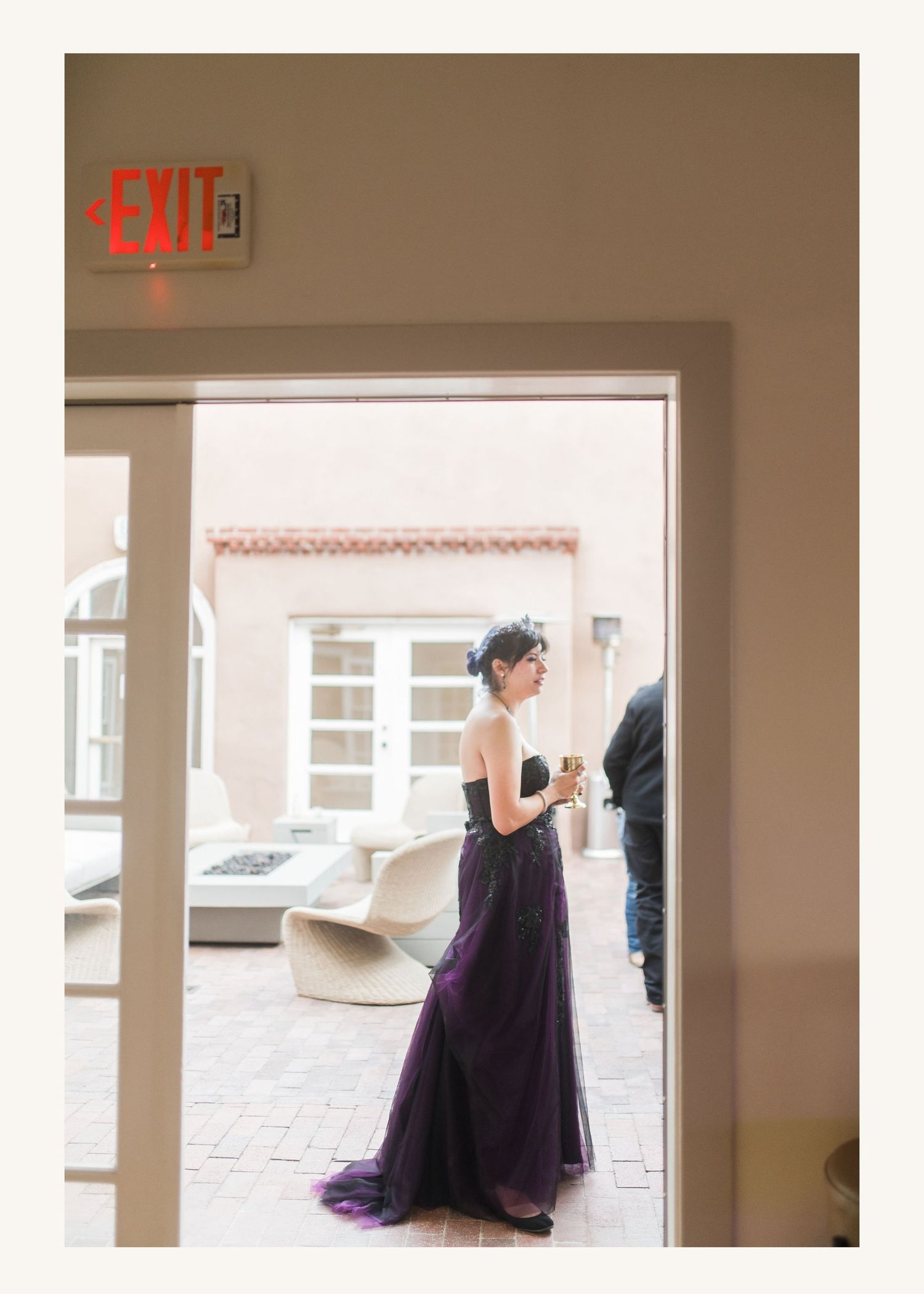Woman in elegant purple strapless gown holding champagne glass on outdoor patio near EXIT sign.