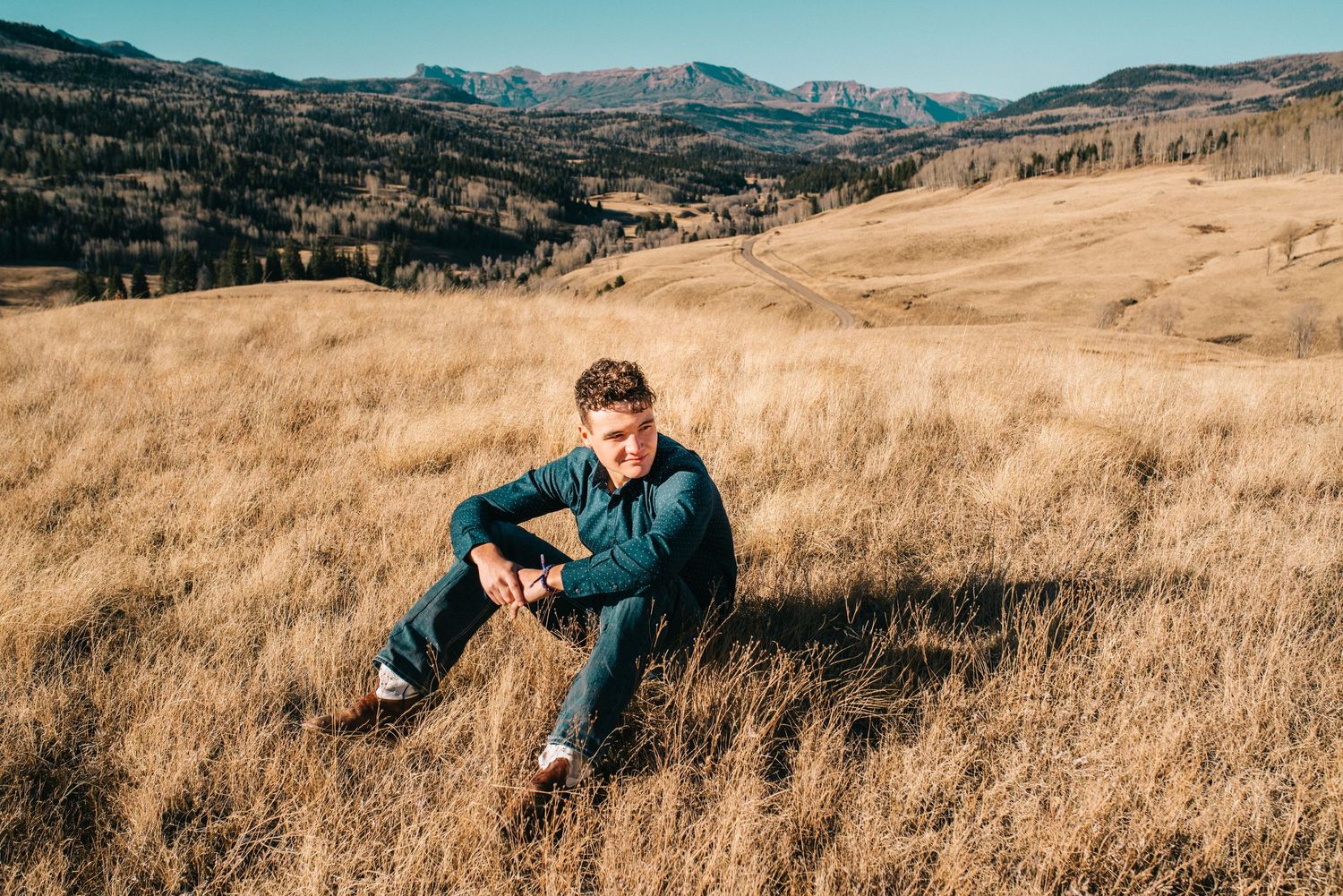Man sitting in golden grassland field with scenic mountain valley backdrop on a clear sunny day.