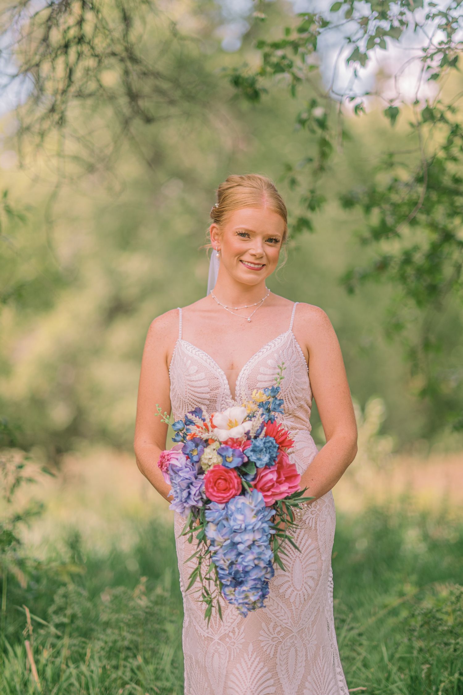 Smiling bride in a beaded gown holds a colorful cascading bouquet with blue, pink, and white flowers outdoors.