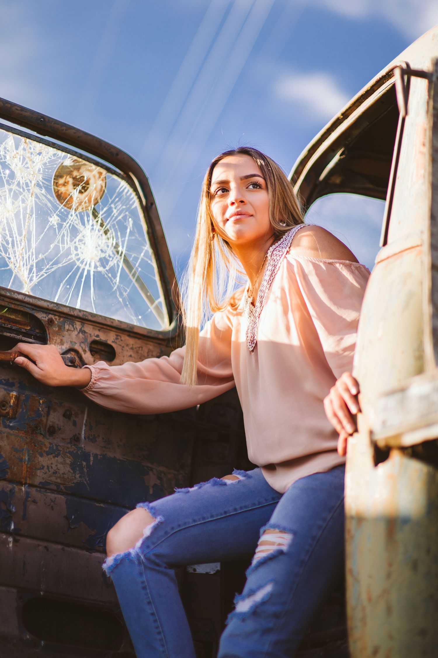 Person sitting in vintage truck with broken windshield wearing pink hoodie and ripped jeans.