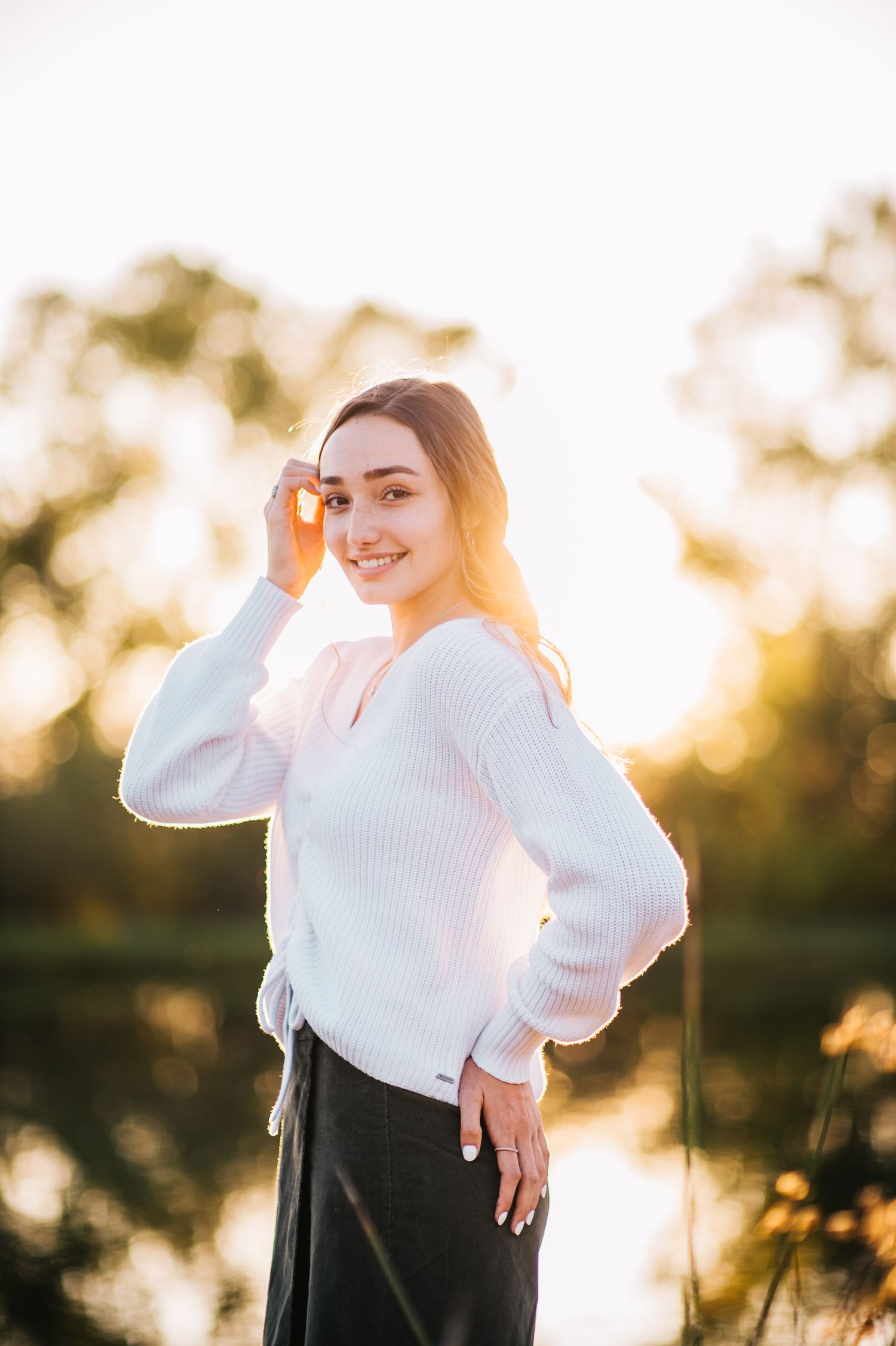 A person in a white sweater poses outdoors during golden hour with trees and sunlight in the background.